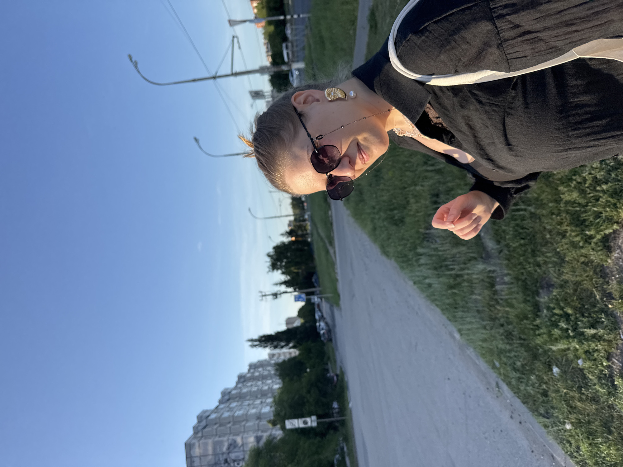 A woman with blonde hair pulled back in a bun, wearing sunglasses, a black collared shirt, and large ornate earrings, stands on the side of a road. She is looking towards the camera with a slight smile. She is also wearing a necklace and has a white bag draped over her shoulder. The background shows a residential building, trees, streetlights, and a blue sky. The image appears to have been taken during the golden hour, with the sun casting a warm glow on the scene.