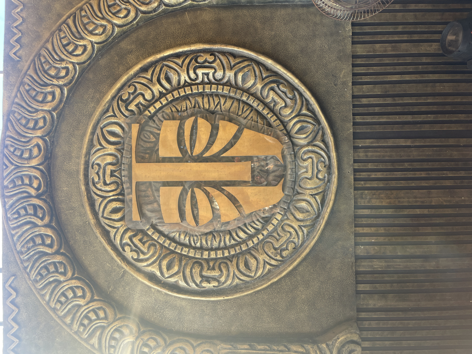 A medium, close-up shot reveals a circular emblem featuring intricate, curvilinear carvings in gold and bronze tones. The emblem is centered within a larger, possibly architectural panel, also finished in a similar gold and bronze color palette. A stylized, geometric design overlays the center of the circular emblem. To the right, the background transitions into parallel lines, possibly metal slats, running horizontally. A partially visible metal fan is also noted at the top of the image. This image was taken in the city of Chopdem, India.