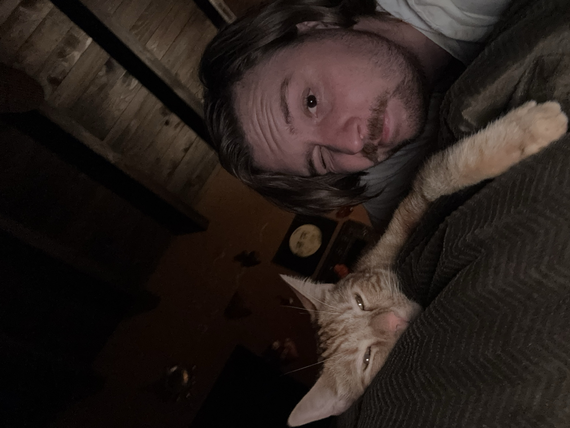 The image depicts a man and an orange cat lying on a dark-colored, possibly corduroy, surface. The man has brown hair and a beard, and is wearing a light-colored shirt. The cat is nestled close to the man. In the background, there is a dark room with a wooden ceiling and a few objects on a table, including a circular item which may be a decorative plate. There is no information available in the image to associate it with the city of Cañada de Madero, Mexico.