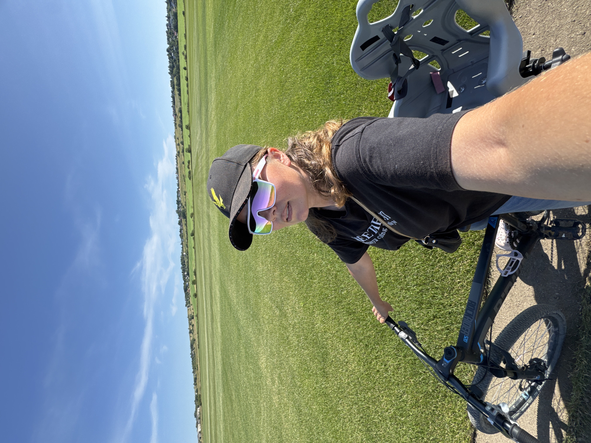 In a grassy field under a clear blue sky, a woman stands on a bicycle, taking a selfie. She wears a black t-shirt, a black baseball cap with a yellow emblem, and white-framed sunglasses with iridescent lenses. The bicycle is black and grey, and a child's car seat is attached to the back. The background shows a vast, manicured green field stretching towards a distant line of trees and houses. The location is identified as Kempelevo, Russia.