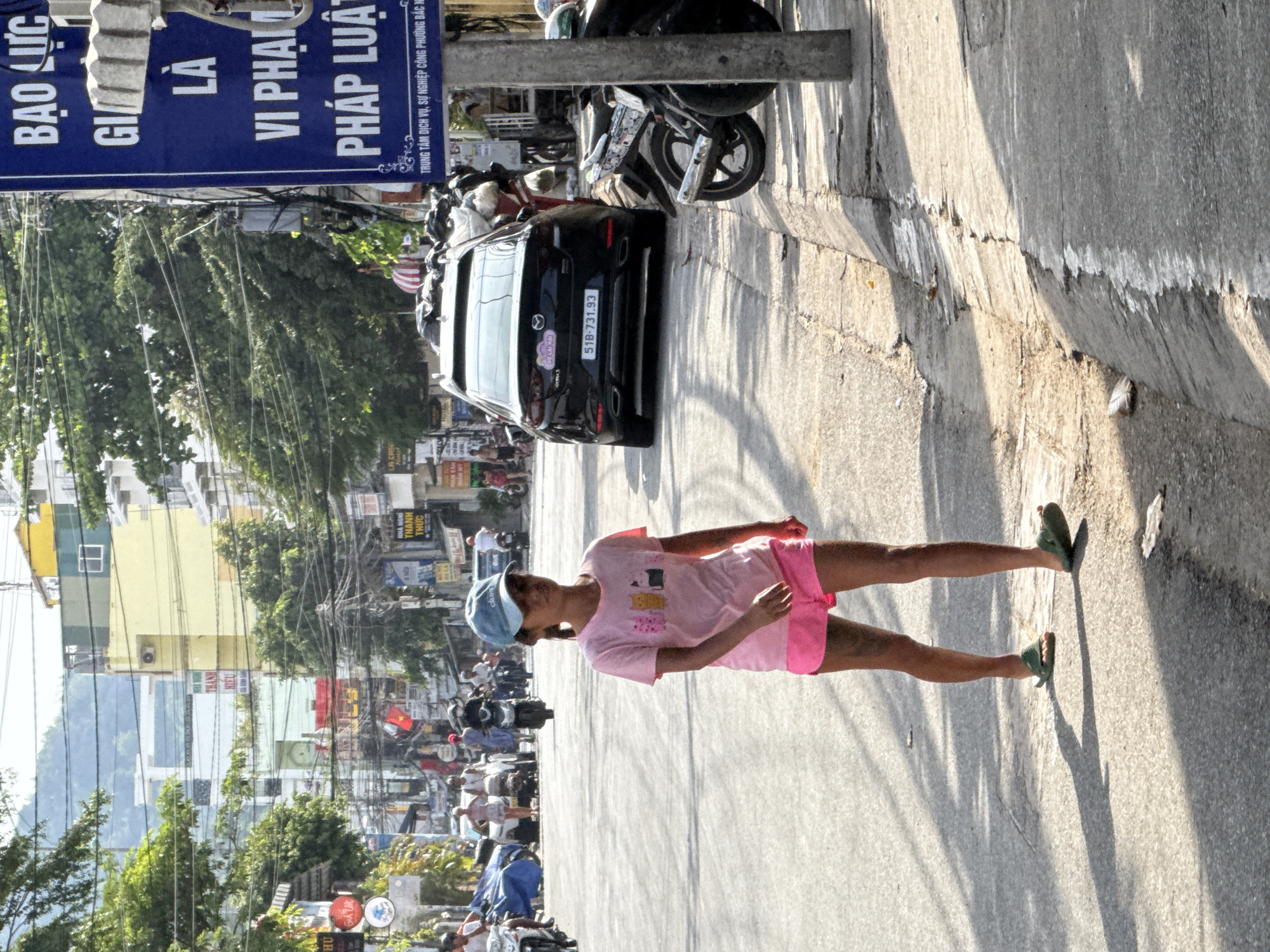 A young girl wearing a pink t-shirt with cartoon cats and pink shorts walks on the right side of a street in Bac Nha Trang Ward, Vietnam. She has on a light blue baseball cap and green flip-flops. Her shadow stretches across the asphalt, which is marked with white lines. In the background, a black Mazda car with license plate "51B-731.93" is parked on the right side of the road, partially obscuring other parked vehicles. A blue sign with white Vietnamese text is affixed to a pole on the far right. The street is lined with trees and buildings of various heights and colors, with power lines crisscrossing overhead. Several motorbikes and other vehicles are visible further down the street, along with pedestrians. The scene is illuminated by bright sunlight, casting long shadows.
