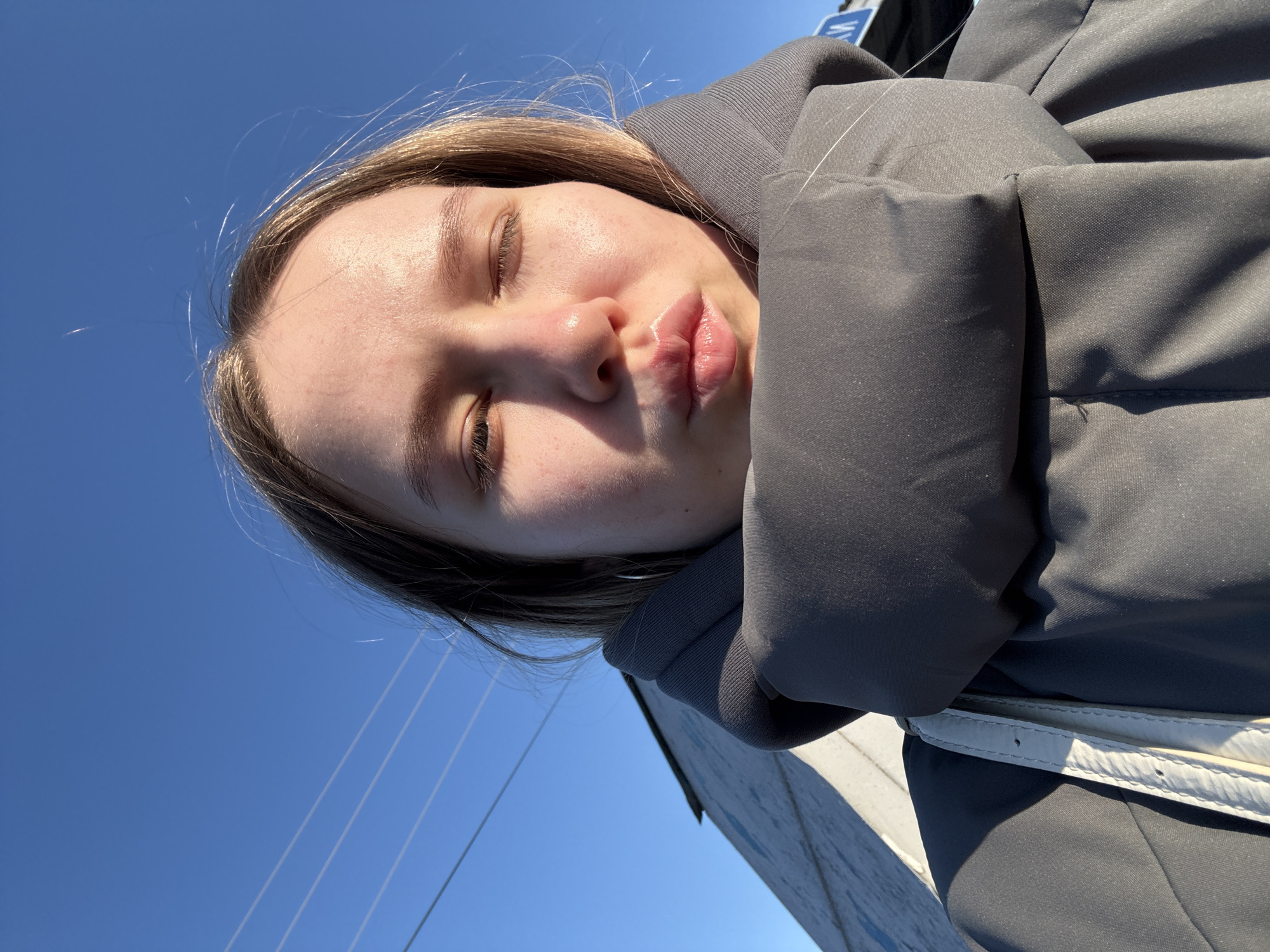 A young woman is pictured in an upward-angle selfie against a clear, bright blue sky. She has fair skin and light brown hair, and wears a gray, hooded jacket. Her eyes are closed, and her lips are pursed. Thin power lines cut across the upper left of the sky. In the lower right corner, a corner of a white building can be seen. The location is Zhigalki, Russia.