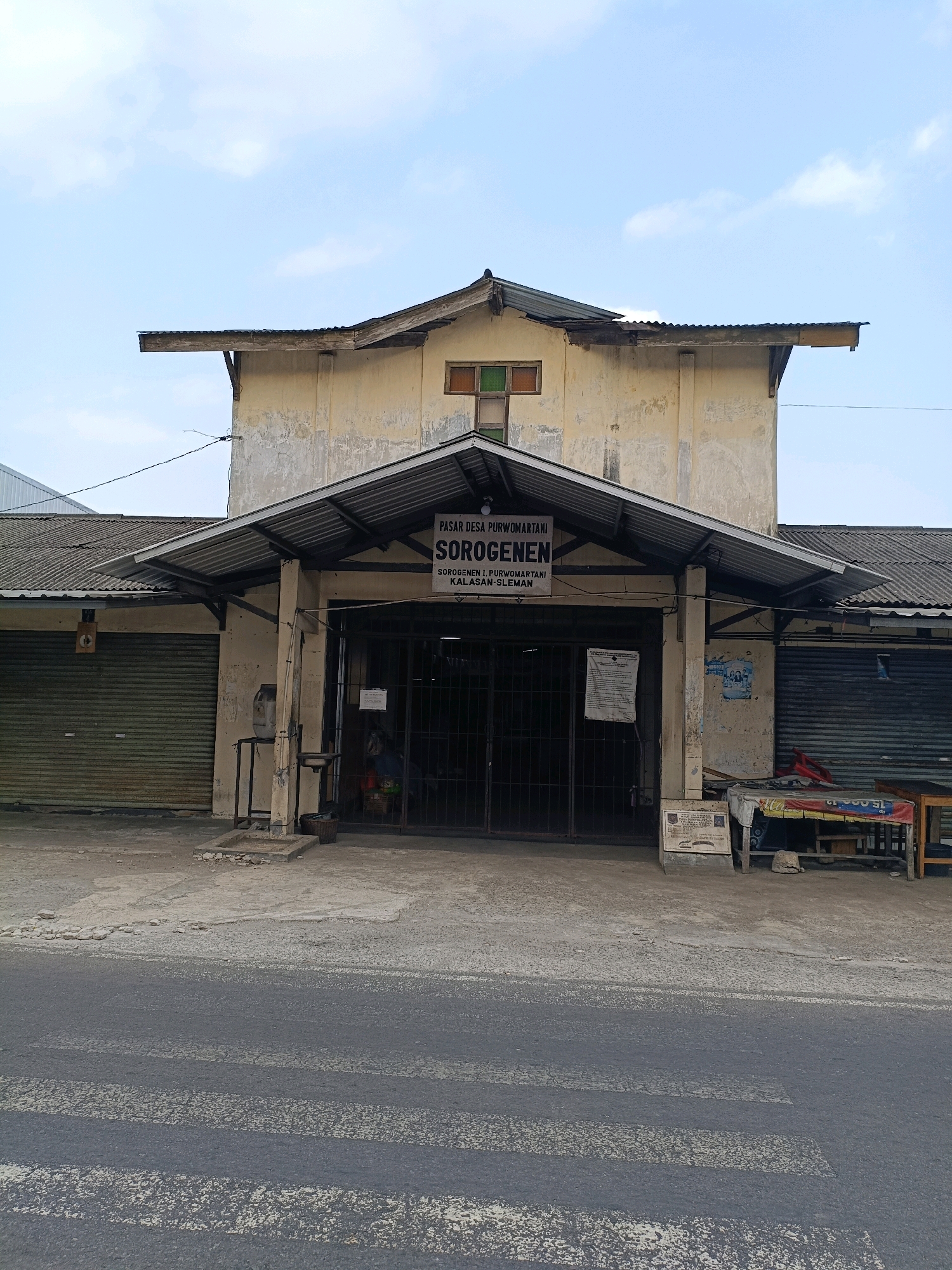 The image depicts the entrance to the "Pasar Desa Purwomartani" (Purwomartani Village Market) in Kalasan, Sleman, Indonesia. The market appears to be a single-story building with a weathered, yellow facade and a corrugated metal roof. The entrance itself is covered by an awning and has a metal gate that is partially open, revealing a dimly lit interior. Signs above the gate indicate the market's name and location. To the left of the entrance, a corrugated metal roll-up door is visible, suggesting a closed stall. To the right, another similar roll-up door is partially visible. The ground in front of the market is paved with concrete and shows signs of wear and tear. A crosswalk is painted on the asphalt road in the foreground. The sky above is partly cloudy. No people are clearly visible in the image, and there are no apparent actions or interactions taking place. The overall impression is of a functional but somewhat aged community market.