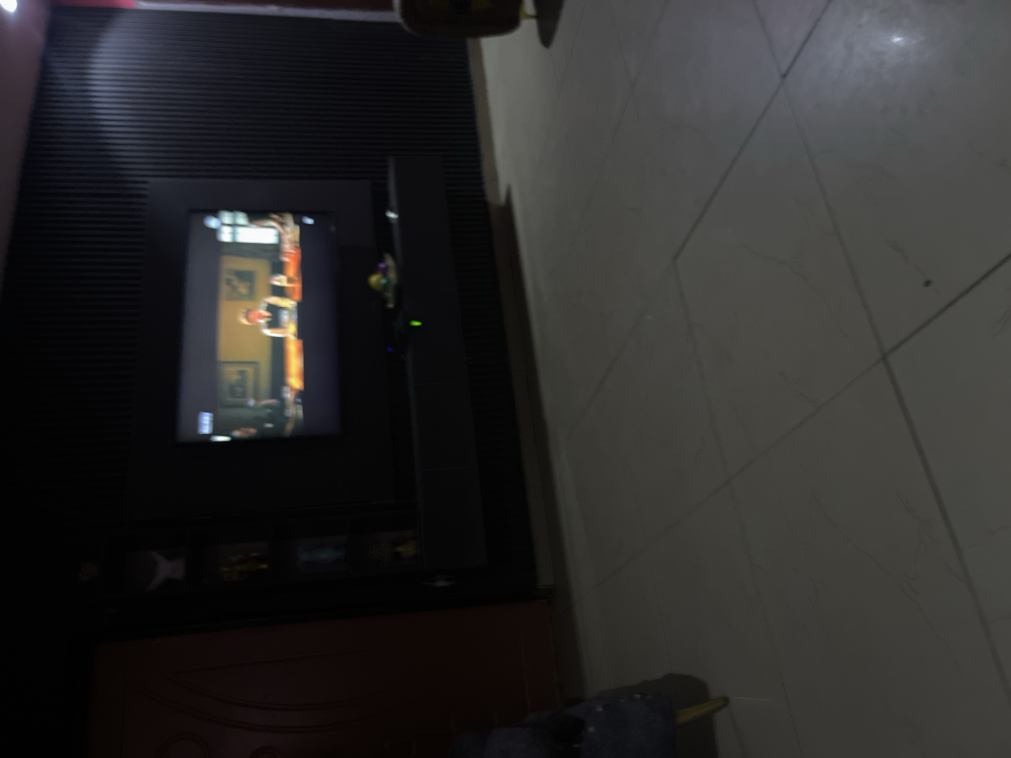 The image depicts an indoor scene, likely a living room, in Jos, Nigeria. A large television screen is mounted on a black, vertically ribbed wall. The screen displays a program with people visible. Below the television is a black floating shelf with some decorative items and a small green light. To the left of the television is a dark door with some decorative elements. A dark colored armchair with golden legs is partially visible in the lower left corner. The floor is tiled with large, light-colored square tiles, some exhibiting a marble-like pattern. The lighting in the room is dim.