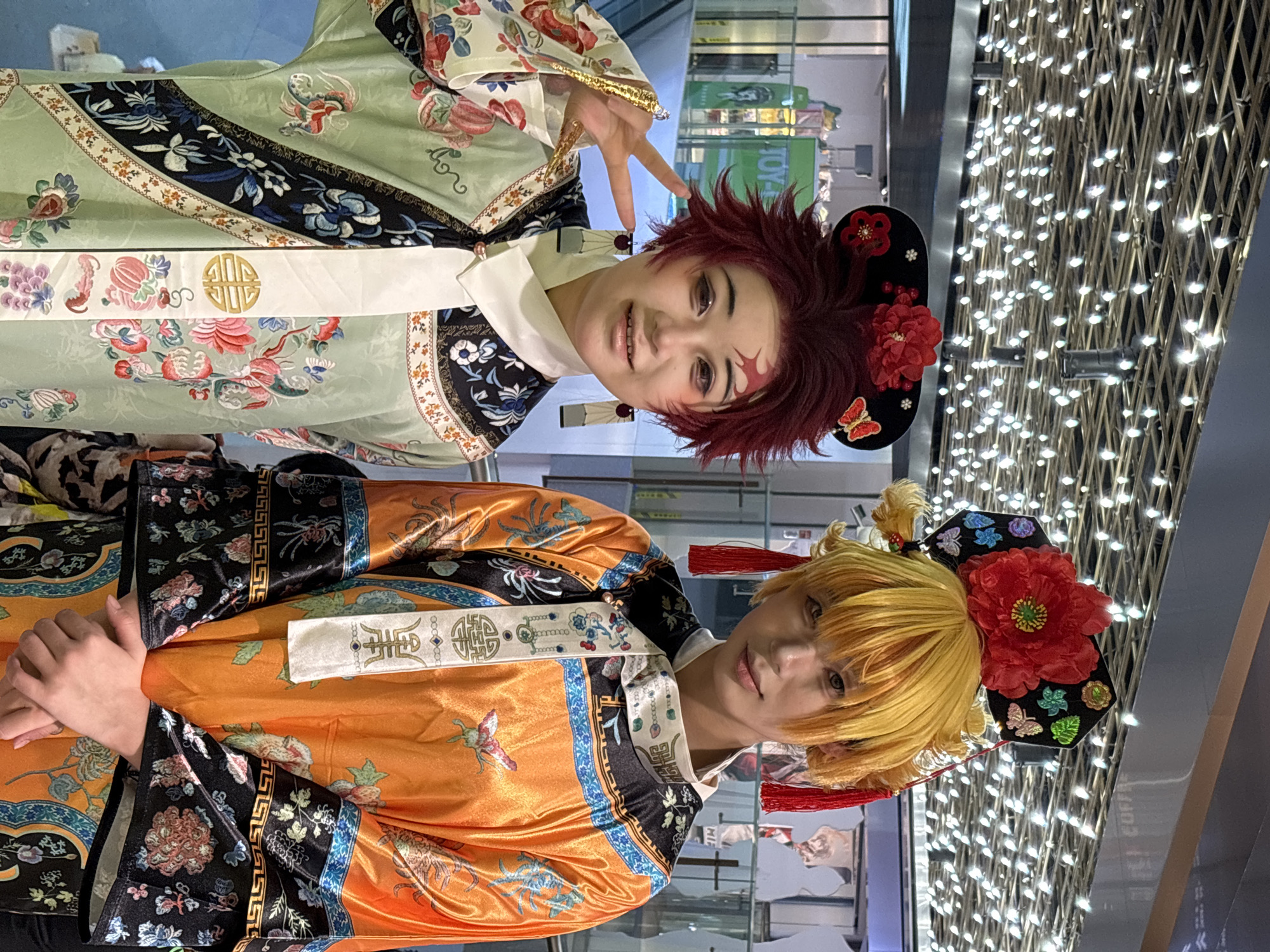 Two individuals in cosplay attire stand indoors. One wears a light green robe with floral patterns and black accents, adorned with a white sash-like belt. The other is dressed in a bright orange robe, also embellished with floral designs and black trim, and a similar white sash. Both have decorative headpieces featuring large red flowers, positioned against a background of illuminated latticework. The setting appears to be in the city of Liangjiang, China, as indicated by the signage in the background.
