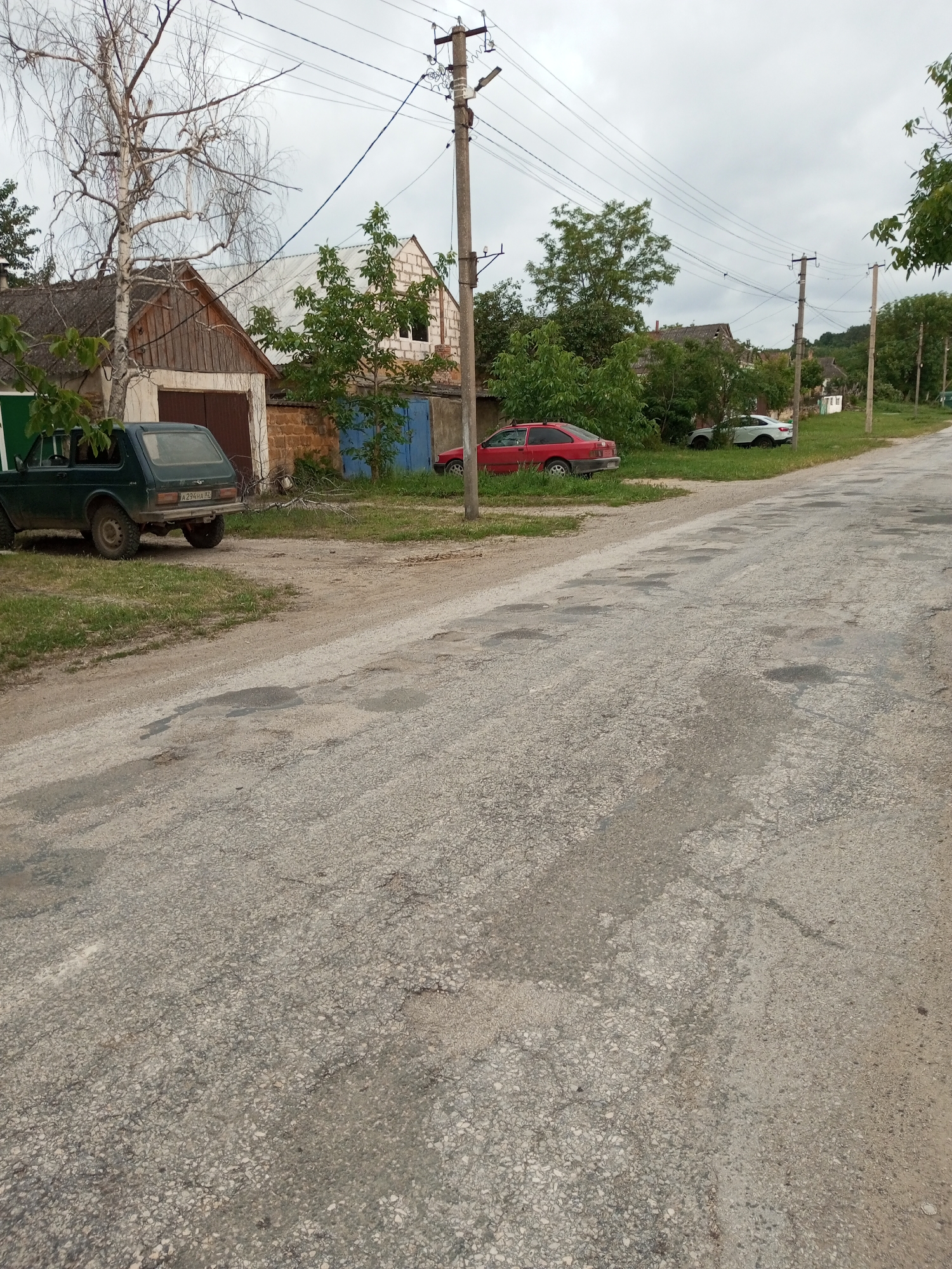 The image depicts a rural street scene in Ароматновское сельское поселение, Ukraine. The foreground is dominated by a cracked and weathered asphalt road with numerous potholes. To the left of the road, a dark green SUV is parked on a patch of grass next to a building with a wooden garage door. Further along the street, a red sedan is parked on the grass, and behind it, a white modern car is also parked. Several utility poles with a network of overhead wires are visible along the road, suggesting it is a populated area. In the background, houses with various roof types, including what appears to be a tile roof and corrugated metal, are scattered amongst trees and vegetation. The sky is overcast, indicating a cloudy day. There are no people visible in the image.