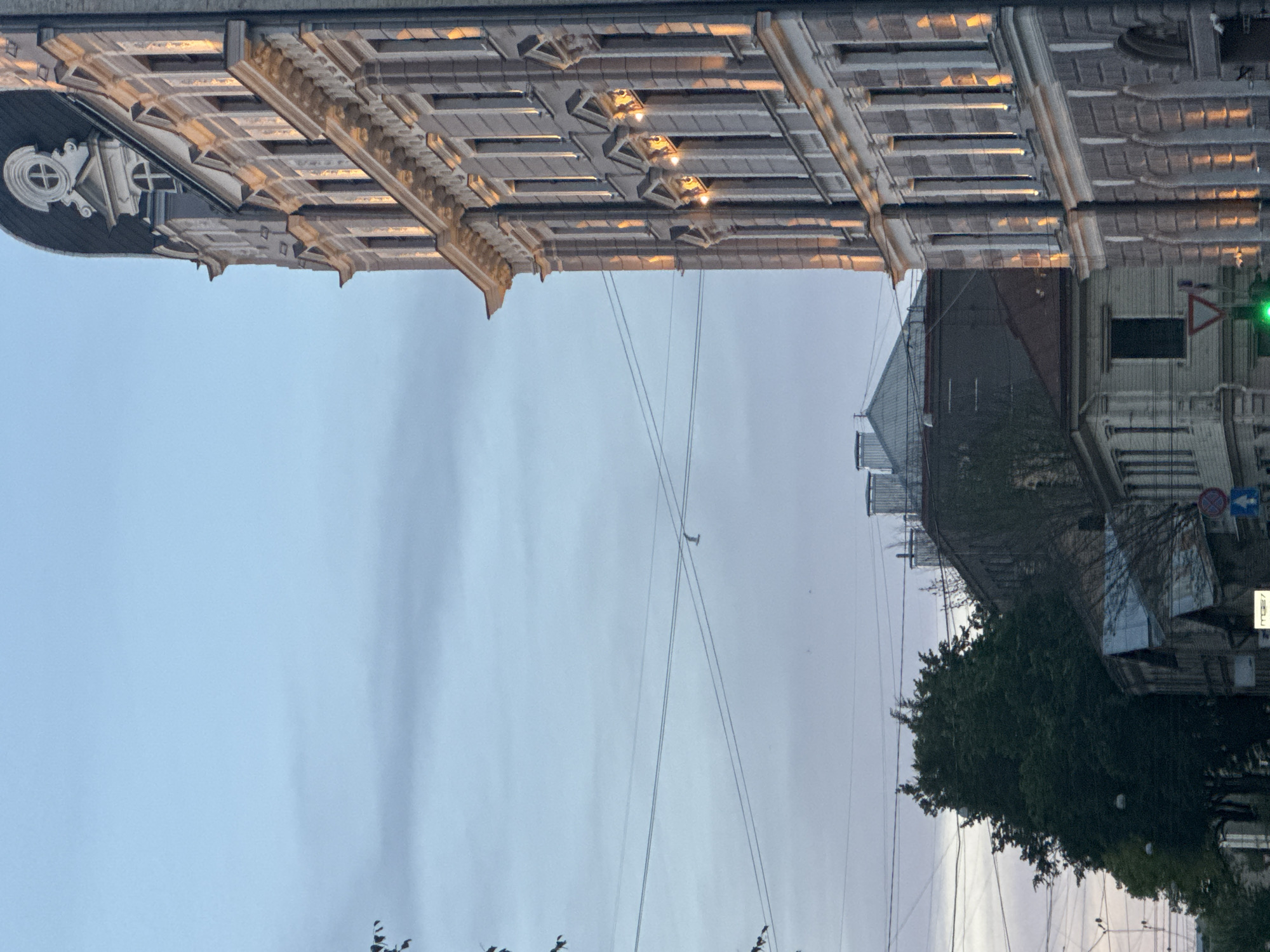 The image shows an exterior view of an old building facade and a street scene in Riga, Latvia. The building's ornate architectural details, including decorative moldings and recessed panels with lighting, are prominent. The sky is overcast, suggesting a cloudy or possibly twilight setting. Below the building, power lines crisscross the sky, a common sight in urban environments.
On the right side of the image, a street is visible with other buildings, trees, and traffic signs. One sign appears to be a circular blue sign with a red border, possibly indicating a restriction, and another triangular sign with a red border and a white interior, which could be a warning sign. A traffic light with a green light is also visible, indicating that traffic is currently permitted to proceed. A tree with dense green foliage stands in the foreground, partially obscuring the view of the street. The overall mood is somewhat subdued due to the overcast sky, but the architectural details of the building add a sense of history and character. No people are visible in the frame. The time of day appears to be either early morning or late evening, given the soft lighting and overcast sky.
