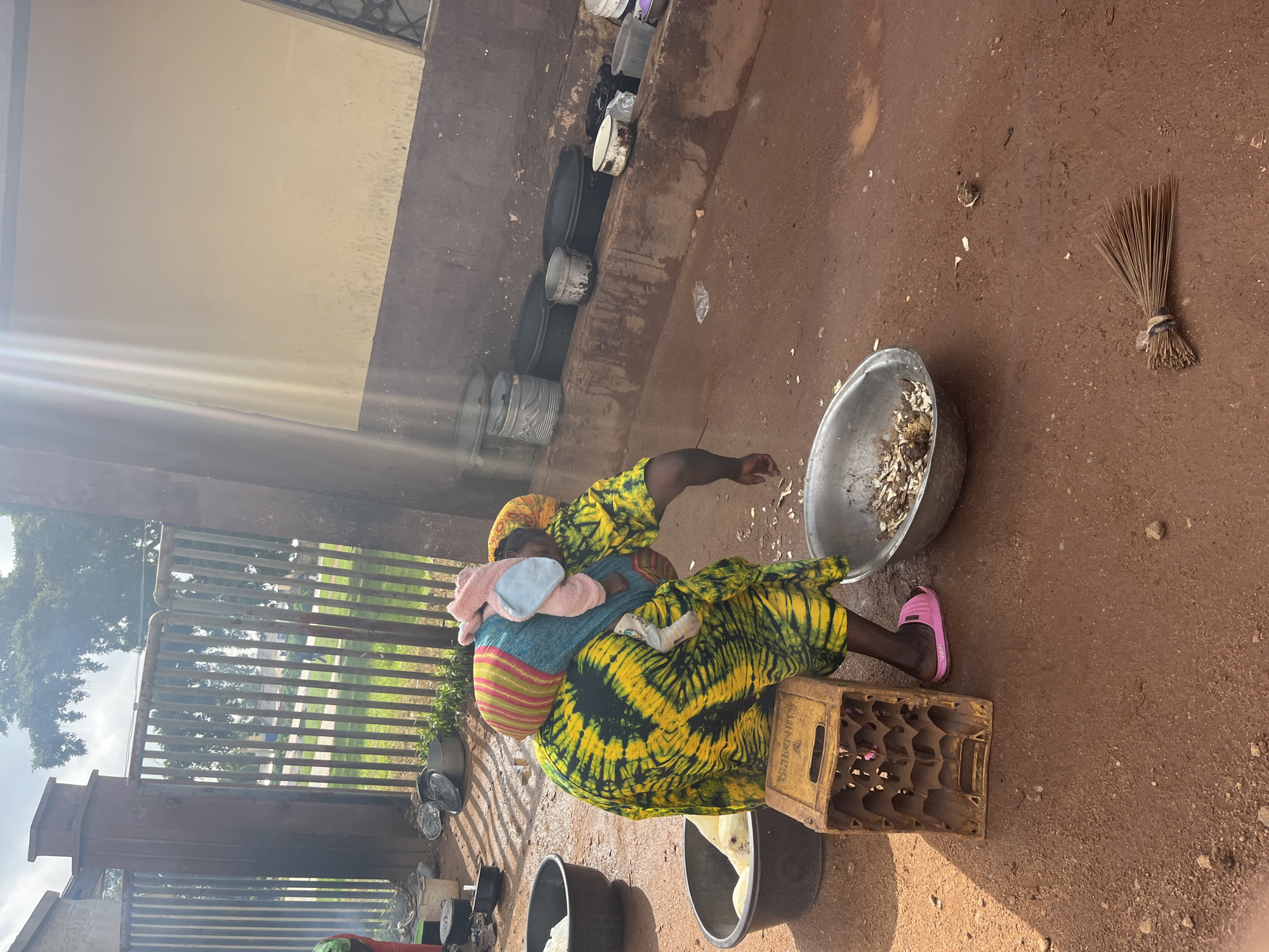 A woman, dressed in a vibrant yellow and green tie-dye patterned dress, is carrying a baby wrapped in a blue cloth and a pink blanket on her back. She is wearing pink sandals and appears to be in an outdoor area with a dirt ground. To her right, there is a large metal basin filled with what looks like food scraps or ingredients. Beside the basin, a wooden crate with the text "GUINNESS" is visible. In the background, there is a fence, some greenery, and a building with a light-colored wall. A broom made of twigs is leaning against the wall to the right of the basin. The scene suggests a simple, possibly rural setting. The lighting indicates it could be daytime, with sunlight casting shadows. The woman's posture and the presence of the baby suggest she might be preparing food or engaged in some daily chores.
