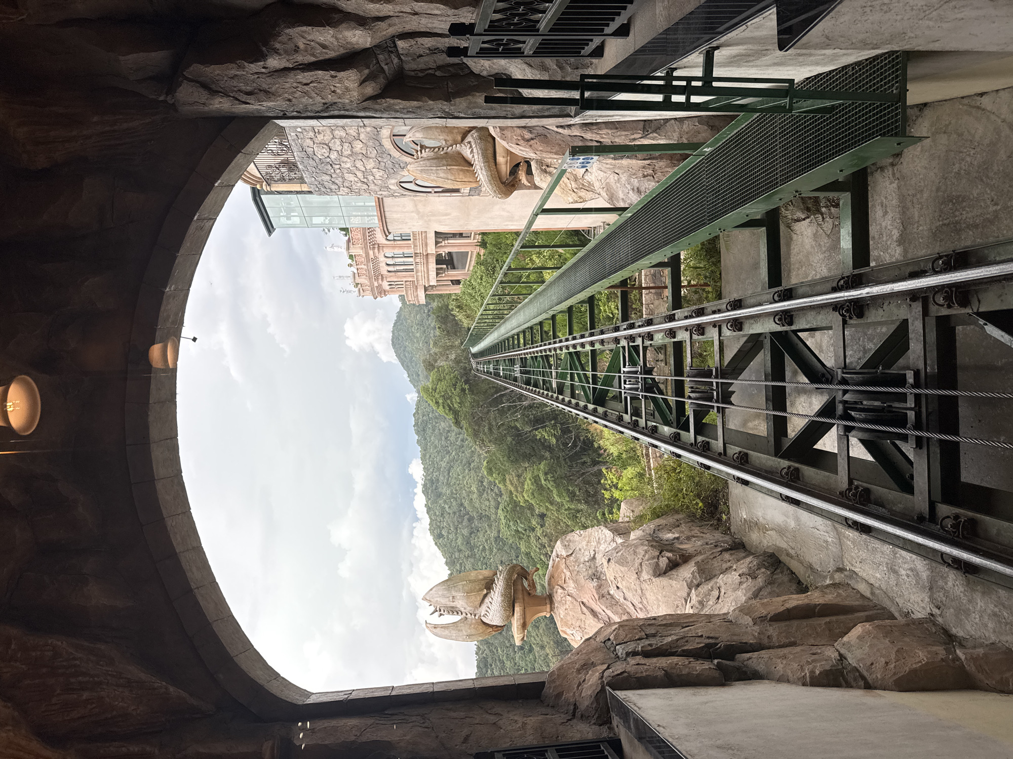 A view through a stone archway reveals a steep funicular railway ascending a mountain. The tracks, supported by a dark metal framework, run directly ahead, drawing the eye upwards towards a dense green forest covering the mountainside. Buildings can be seen partially obscured by the archway and foliage. Stone dragon sculptures adorn the sides of the passage. The scene is likely located in the Ba Na Hills of Đà Nẵng, Vietnam, a mountain resort known for its unique architecture and cable car systems.