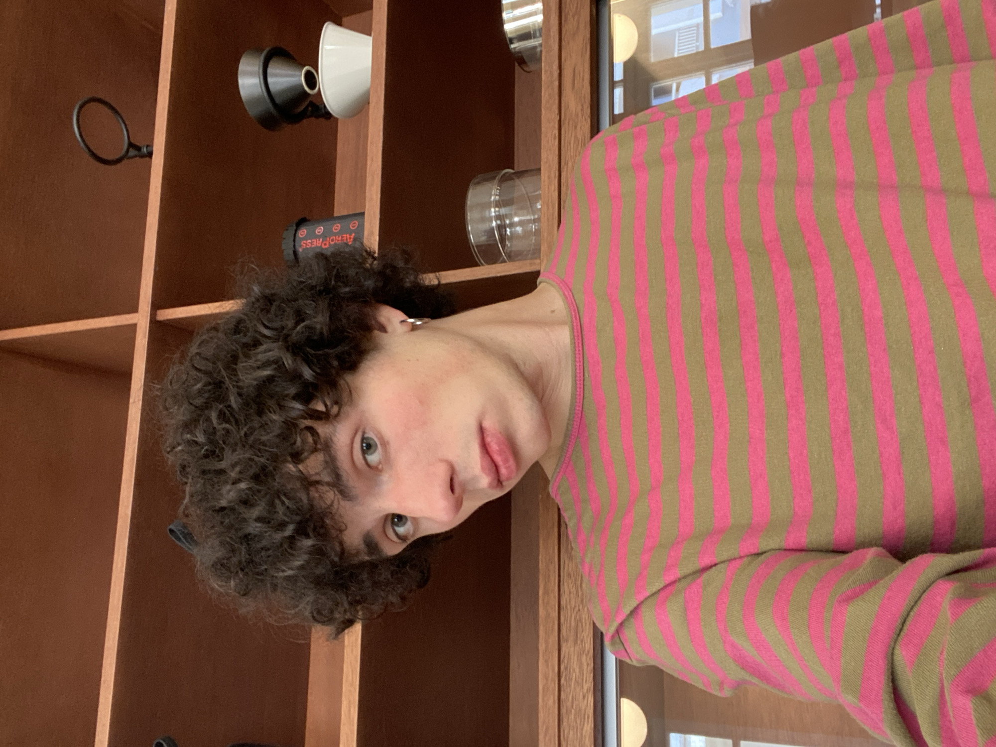 A portrait shot depicts a young, fair-skinned individual with tightly curled, dark brown hair and blue eyes. The person is wearing a striped shirt in pink and olive-green. The background features a wooden shelving unit displaying coffee-making apparatus like a pour-over coffee maker, an Aeropress, and various glass containers. The presence of an overhead light suggests an indoor setting, potentially in Tel Aviv, Israel. The specific location within the city is not discernible from the image.