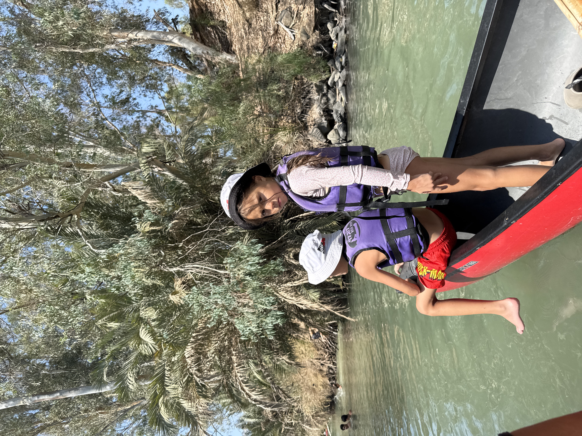 Two children are in a red canoe on a river in the Emek HaYarden Regional Council, Israel. The child in the foreground is wearing a purple life vest, red shorts, and a white bucket hat. The child behind them is wearing a purple life vest and a patterned swimsuit. They are surrounded by lush vegetation and trees on the riverbank. The water is a murky green color. In the background, other people are swimming in the river.