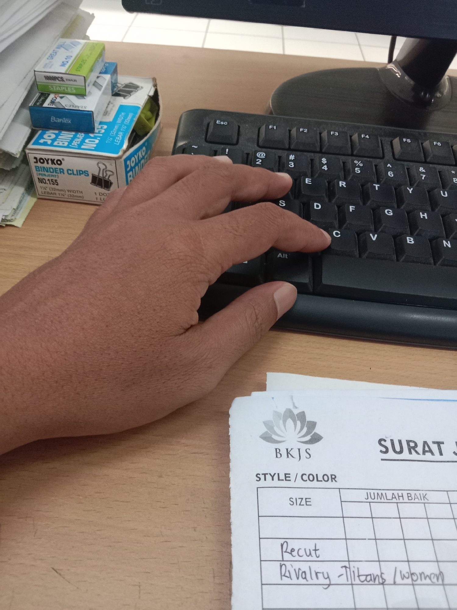 A person's hand is resting on a keyboard, with their index finger pressing down on the 'C' key. In the background, a stack of stationery items, including boxes of binder clips and staples, are visible on a wooden desk. A document with the heading "SURAT J" and fields for "STYLE/COLOR", "SIZE", and "JUMLAH BAIK" is also present, with handwritten entries "Recut" and "Rivalry-Titans / women". The scene appears to be an office environment. The image does not contain enough information to determine a specific location in Semarang, Indonesia.