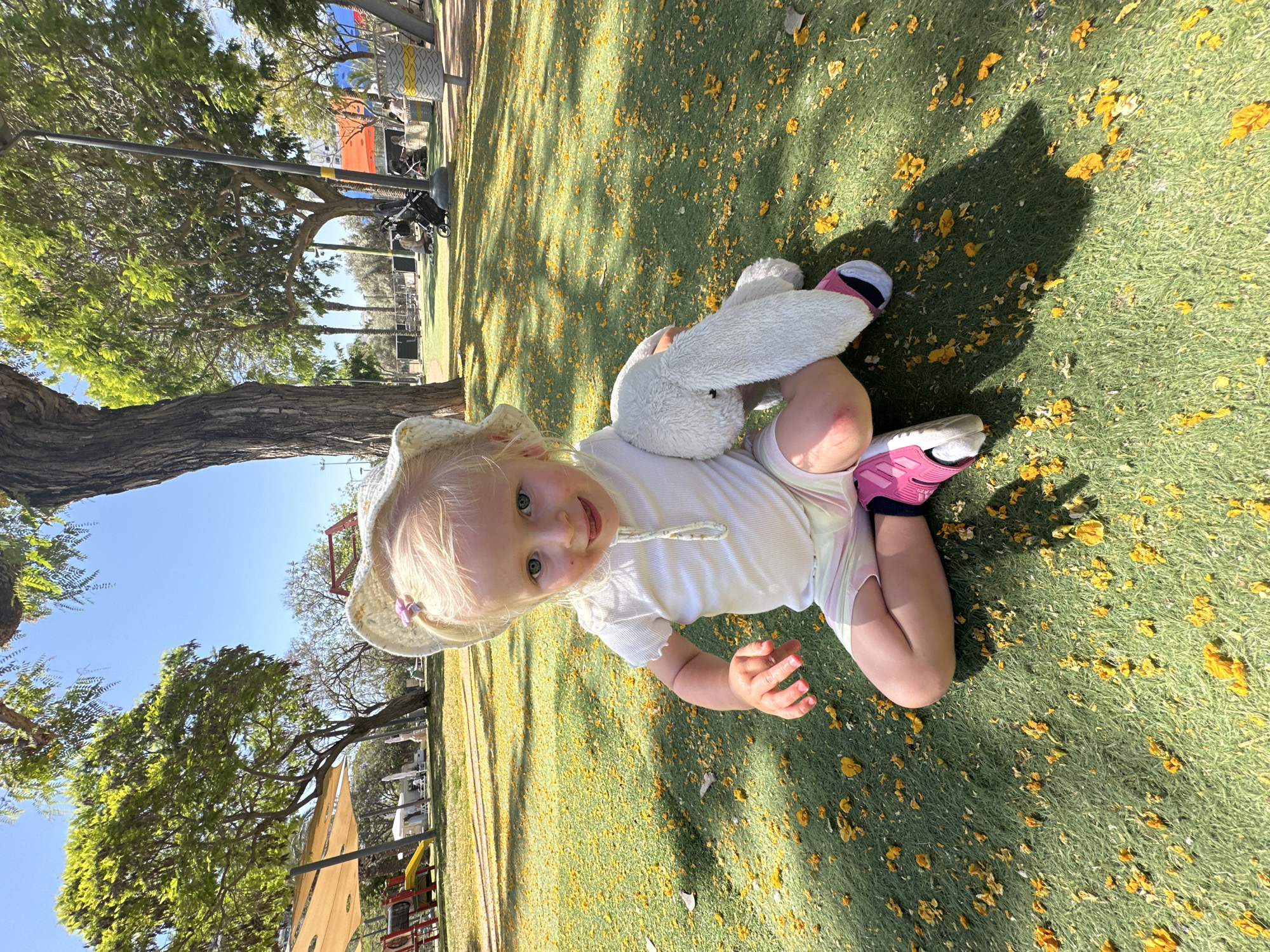 This image depicts an outdoor scene in a park or playground located in Petah Tikva, Israel, under a clear blue sky on a sunny day. The central subject is a young child, likely a toddler, with blonde hair, wearing a white ribbed short-sleeved top, light-colored striped shorts, a white sun hat with a pink flower clip, and pink and white athletic shoes. The child is seated on green artificial turf, which is densely scattered with bright yellow fallen flowers or petals. The child faces the camera with a slight smile, holding a white, fluffy stuffed bunny rabbit in the left arm. Red marks are visible on the child's right cheek and left knee.
In the background, a large, mature tree with green foliage is prominent on the left, with its trunk forming a vertical element. Further to the left, under a large beige canopy, red and yellow playground equipment, including a slide, is discernible. Various other trees with green foliage are scattered across the mid-ground and background. To the right of the child, a dark grey light pole extends vertically. Further back on the right, a yellow and grey patterned cylindrical object, possibly a trash can or bollard, is visible. Several adults and at least one baby stroller are present in the mid-ground, indicating other park visitors. The sunlight casts distinct shadows on the green turf, originating from the child and other objects in the scene.