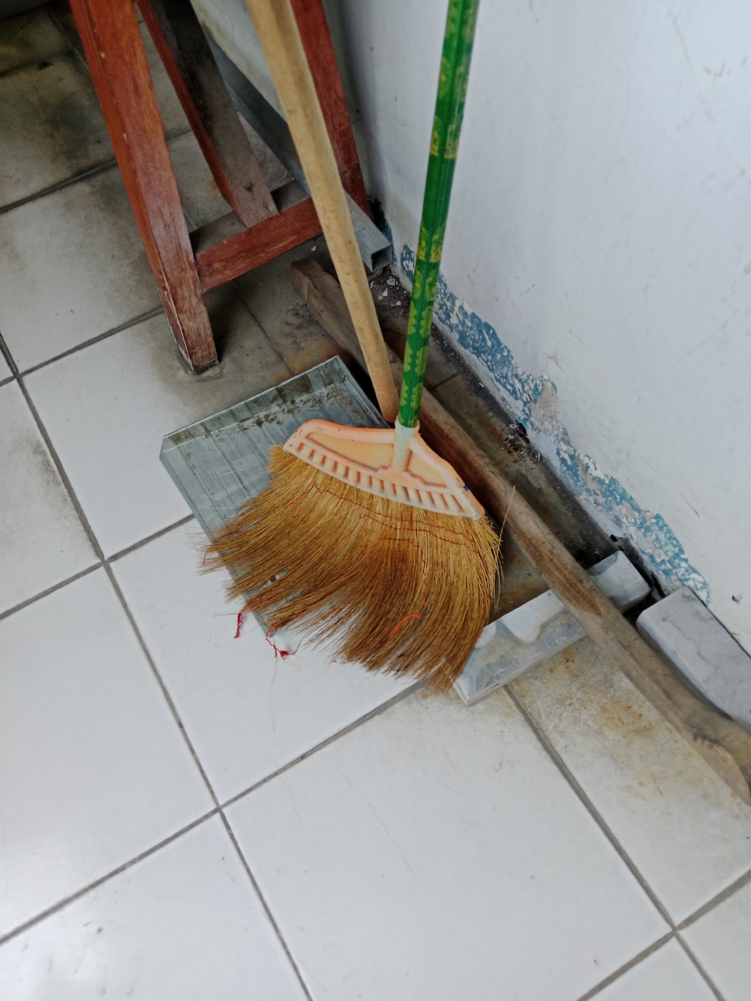 The image displays a broom with a green and yellow patterned handle resting on a dustpan, which is positioned on a white tiled floor. A wooden ladder and another broom with a light-colored handle are also visible. The scene appears to be an indoor space, possibly a corner of a room, with a white wall and a blue-stained baseboard. There are no people or animals present in the image. The overall impression is that of a cleaning area or storage space. The objects suggest a domestic or utilitarian setting.