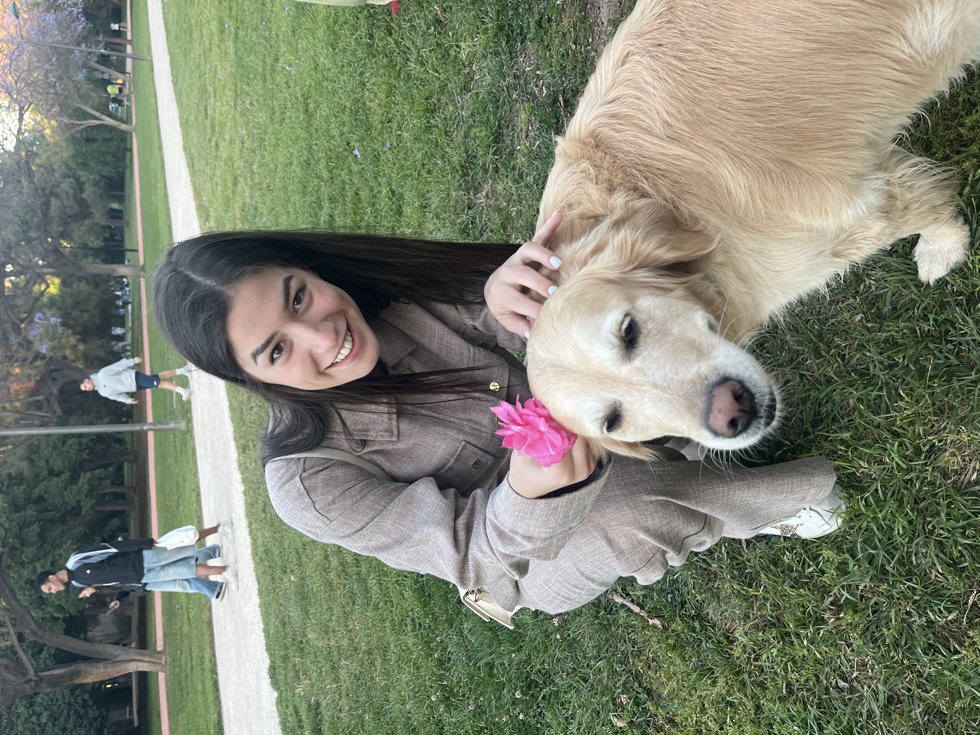 A young woman and a golden retriever dog are sitting on grass in a park. The woman is wearing a brown jacket and pants, and she is holding a pink flower. The dog is lying down next to her, with its head resting on her lap. In the background, there is a paved pathway with two people walking on it. The park appears to be located in the city of València, Spain.