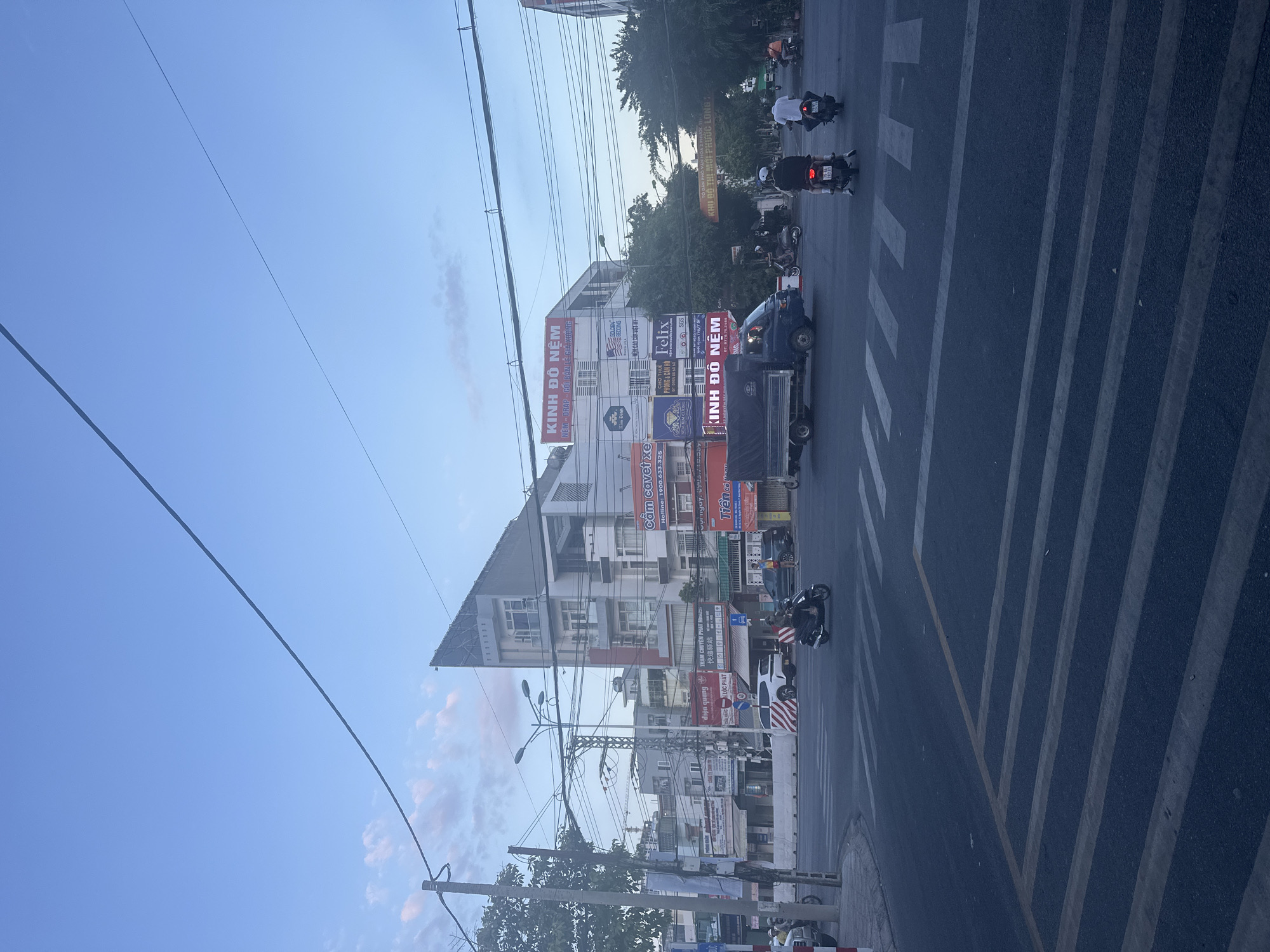 The image depicts a street scene at dusk in Nam Nha Trang Ward, Vietnam. The sky is a pale blue with wispy clouds. Several power lines crisscross the frame. In the foreground, a paved road features white pedestrian crosswalks. A dark blue truck with a covered cargo bed is driving through an intersection. Multiple motorcycles are also present, with riders visible on some. One motorcycle is being ridden by a person wearing a white helmet, and another by two individuals, one wearing a white shirt. Buildings line the street, many with signage in Vietnamese. One prominent building has a uniquely angled roof. Trees with dense foliage are visible on the right side of the image. The overall impression is of an active urban intersection in the early evening.