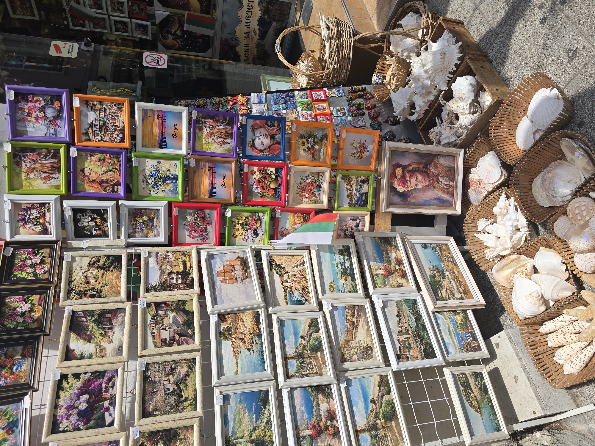 The image depicts a street stall in Burgas, Bulgaria, displaying an assortment of framed artworks and decorative shells. The artworks are predominantly landscape paintings, featuring scenes of coastal towns, seascapes, and floral arrangements, many with colorful frames. Some paintings are labeled with "Burgas," confirming the location. In the foreground, several woven baskets overflow with an array of seashells and starfish, arranged for sale. Behind the stall, a shop window is visible, displaying more items and bearing Bulgarian flag imagery. The overall scene suggests a tourist-oriented market or shop.