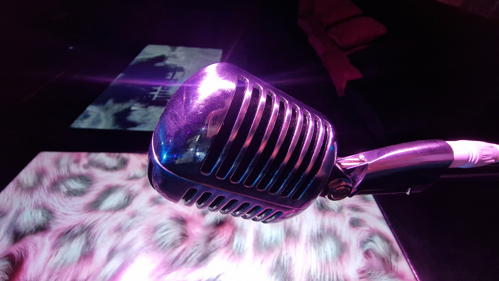 A close-up shot focuses on a vintage-style microphone, brightly illuminated with purple and blue lighting. The microphone's head is chrome with vertical bars. The connector is a pinkish-purple. In the blurred background, a screen displays abstract images. One screen shows a monochromatic scene, while another closer to the microphone, features a pink and white animal print-like pattern. Further in the background there is a black couch with a red throw blanket. The location is likely a studio or performance space in Baan Bang Rak, Thailand, although there is no explicit confirmation of this detail in the image.