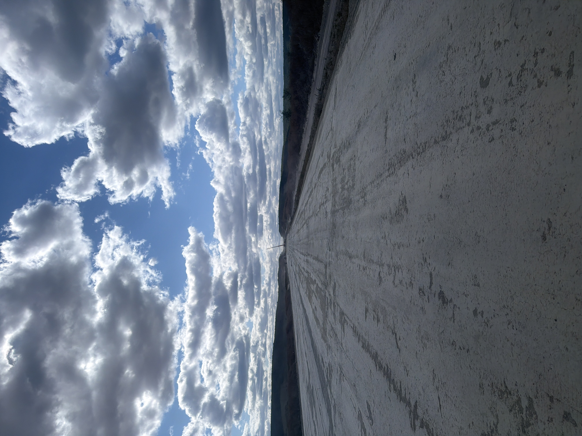 The image depicts a vast, desolate, and unfinished road or construction site stretching into the distance. The ground surface is a light-colored, compacted material, likely dirt or gravel, showing tire tracks and uneven textures. The road is flanked by low, dark, tree-covered hills on either side, creating a sense of a rural or undeveloped landscape. In the far distance, a tall construction crane is visible, suggesting ongoing development or infrastructure work. The sky above is dramatic, filled with a mix of dark, heavy clouds and bright patches of blue sky, indicating a partly cloudy day. The overall impression is one of open space and a work in progress, set against a natural backdrop. The location of Колотов Буерак, Russia, is context for this scene.