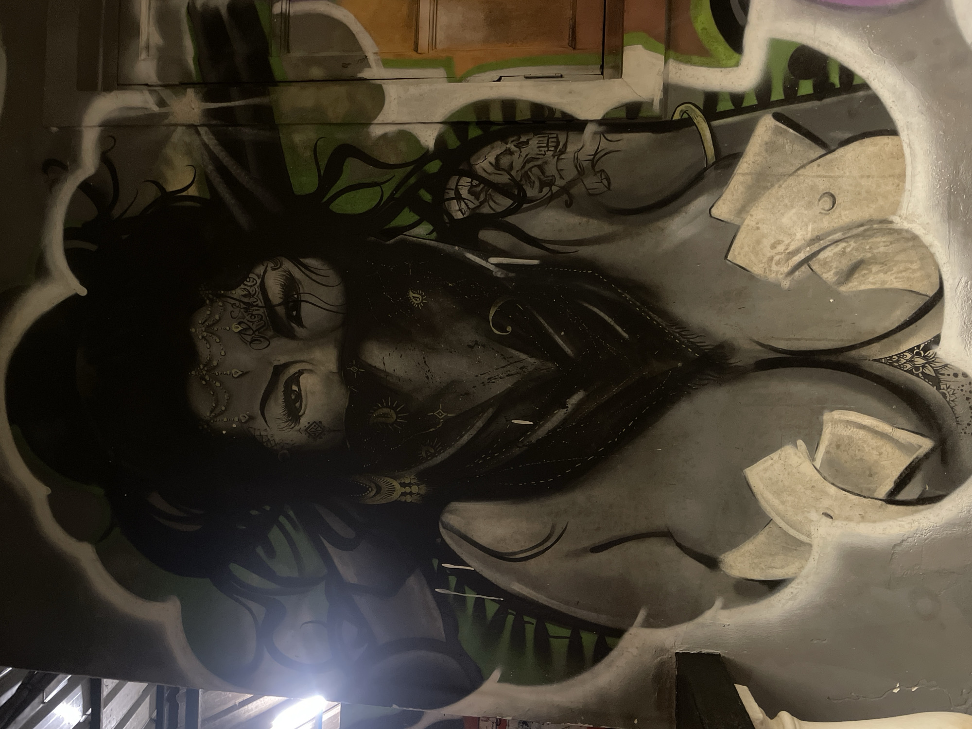 The image displays a graffiti mural on a wall in Vagator, India. The mural features a stylized portrait of a woman with dark, wavy hair and a bandana covering her lower face. She has prominent eyes, elaborate jewelry on her forehead and ears, and a tattoo of a skull on her right arm. Her bare chest is depicted with large, rounded breasts, and her hands hold what appear to be folded bills, possibly money. The background of the mural is abstract, with swirling gray and green elements. The overall style is illustrative and somewhat dramatic. The mural is painted on an outdoor wall, and the surrounding environment is dimly lit, suggesting it is nighttime or in a shaded area.