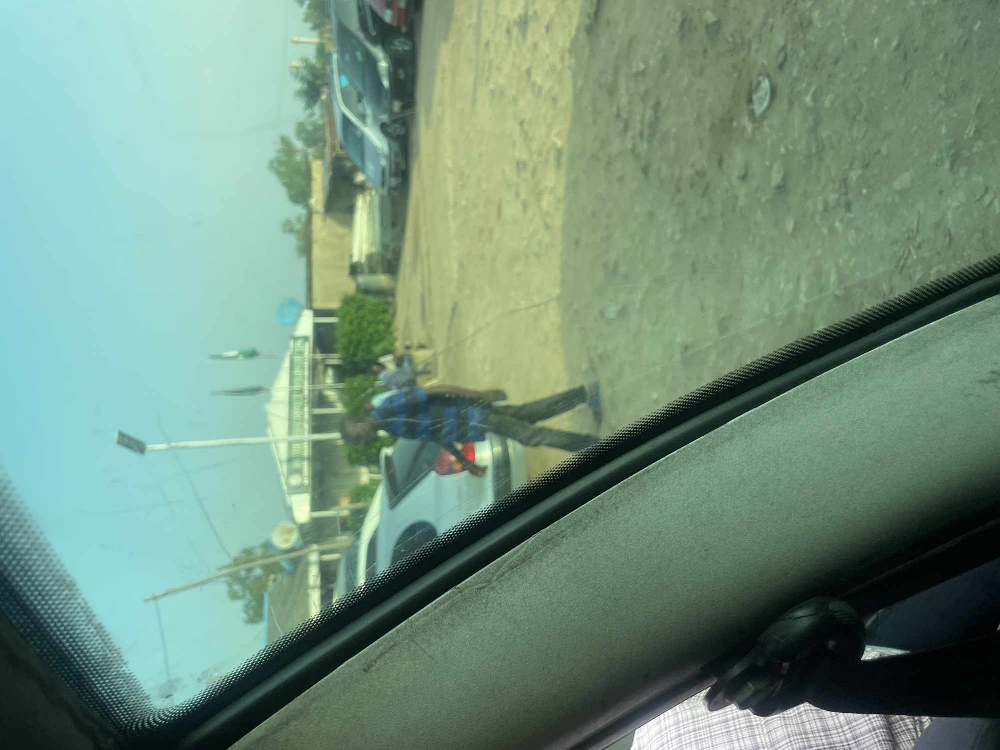 The image is a photograph taken from inside a vehicle, looking out through the windshield and a sunroof. The perspective is distorted and the image is rotated. The scene appears to be a dusty, outdoor environment with several vehicles and buildings visible. The time of day seems to be daylight, with a bright, hazy sky.
In the center of the frame, a person wearing a blue and black checkered shirt and dark pants is walking. They appear to be walking away from the camera and towards a light-colored car. The ground is a light brown, dusty dirt or gravel. In the background, there are several other vehicles, including what looks like a white van or SUV, and other cars that are partially obscured. Buildings with simple architecture are also visible, some with corrugated metal roofs. There are some trees and foliage scattered throughout the scene, suggesting it might be a somewhat rural or semi-urban area.
The overall impression is of a typical day in a place with unpaved roads and a moderate amount of vehicle traffic. There are no discernible emotions conveyed by the subjects, and no visible text in the image. The quality of the image is somewhat grainy, likely due to the lighting conditions and the angle of the shot. The windshield and sunroof of the vehicle from which the photo is taken are also visible, framing the outdoor scene.