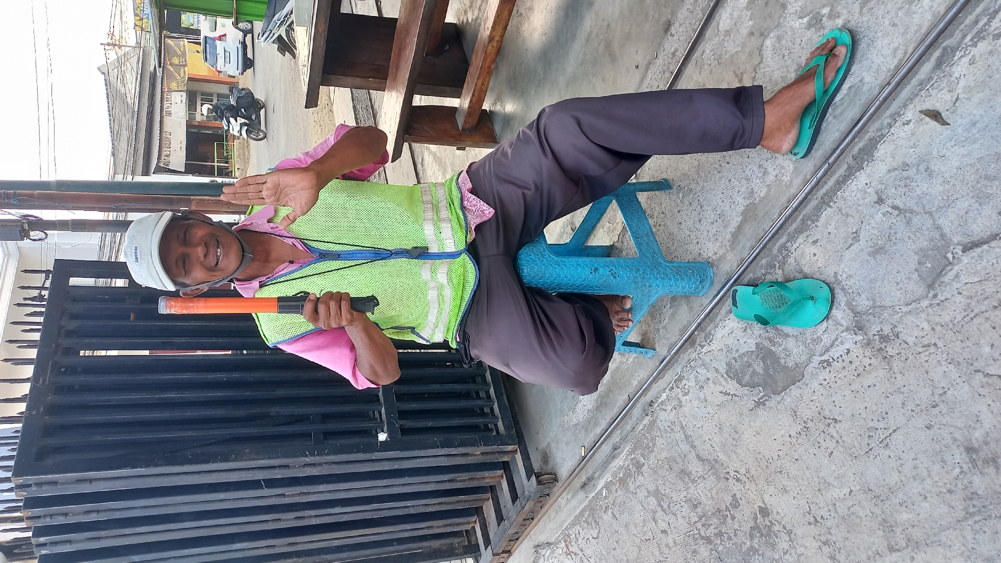 A man wearing a white baseball cap, a pink shirt, and a neon green safety vest sits on a blue plastic stool in front of a black metal gate. He holds an orange and black traffic wand in his right hand and raises his left hand in a greeting gesture. His pants are dark gray, and he is wearing green flip-flops. The ground is paved with concrete, and a metal track runs along the foreground. In the background, there are buildings, a wooden bench, and vehicles on a street. The scene appears to be in a street or an intersection in Mangunsari, Indonesia.