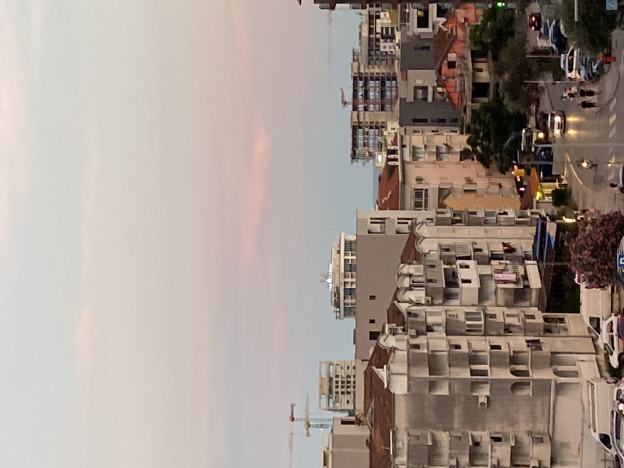 The image captures an urban cityscape at dusk in Budva, Montenegro. The sky is a soft gradient of pink and pale blue, with a hint of pink cloud formation in the upper center. In the foreground, a cluster of apartment buildings with distinctive balconies and architectural details dominates the left and center of the frame. Laundry hangs from some of the balconies.
Further into the city, taller buildings are visible, including a distinctive circular building with the word "THE CANNE" illuminated on its rooftop. Construction cranes are present in the middle ground, indicating ongoing development. The middle ground also features a mix of residential and commercial buildings, with some appearing to be older structures with red-tiled roofs.
A street runs through the lower right portion of the image, with several cars and a motorcycle visible. People are walking on the sidewalk. The streetlights and vehicle headlights are beginning to illuminate the scene, reflecting on the wet pavement, suggesting recent rain or the aftermath of a shower. The overall impression is of a vibrant, developing coastal city transitioning into evening.