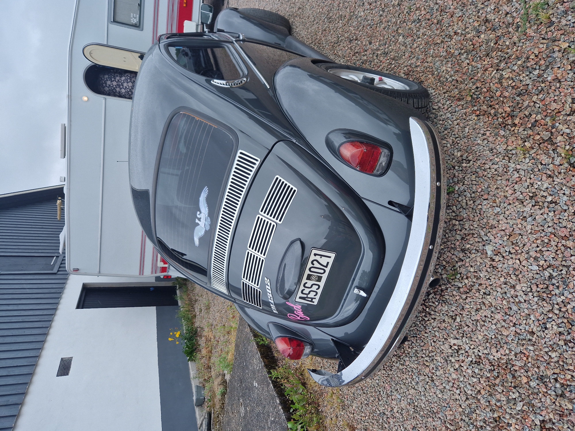 The image shows the rear of a dark grey Volkswagen Beetle parked on a gravel surface. The car has a chrome bumper and a license plate that reads "ASS 021". There is a pink sticker above the license plate with the word "Bad" written on it. A white bird decal is visible on the rear window. In the background, a white caravan is parked next to a building with a dark grey wooden facade and a white wall. The scene is set in the city of Vessigebro, Sweden.