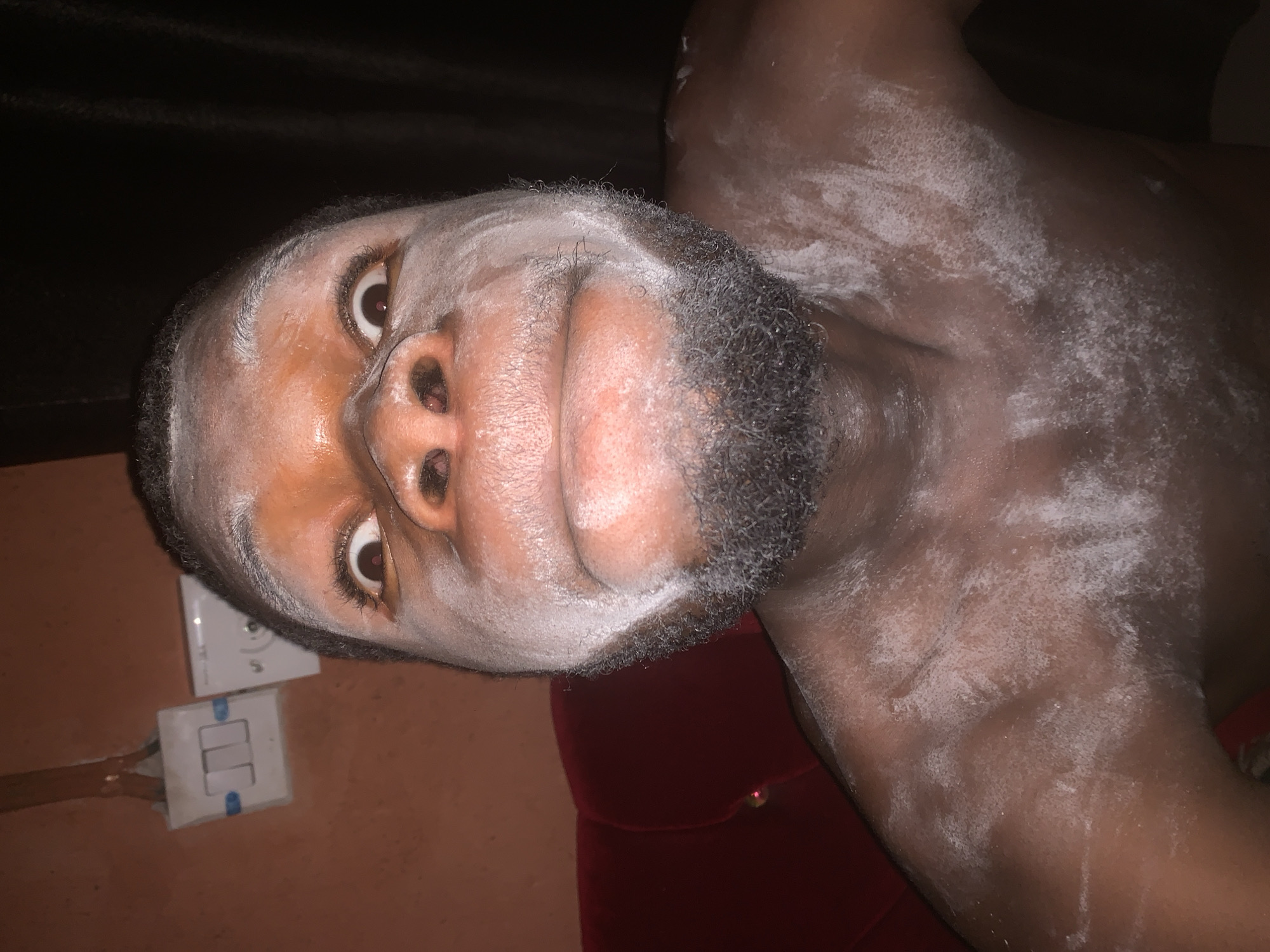 A dark-skinned man is depicted from a rotated perspective, appearing to lie on his back and look towards the camera. His face and upper torso are largely covered in a white, powdery substance that contrasts sharply with his skin tone. The powder is visible across his forehead, cheeks, nose, and extends onto his beard and upper chest/shoulder area. His eyes are wide open, gazing directly towards the viewer, with dark irises and visible whites. He has a dark, well-groomed beard.
The activity involves the application of this white substance, which could be chalk, cosmetic powder, or part of a cultural/ritualistic practice. The man's expression is attentive, with a slight, subtle smile on his lips.
The scene appears to be indoors, likely a room, with a dark, indistinct background above and to his right. To his left and below him, a light orange-brown wall is visible, featuring two white electrical switches or outlets, one positioned above the other. The lower switch has two toggles, one of which appears to be blue. Some wiring is visible near these fixtures. A dark red, possibly velvet, cushion or piece of furniture is seen partially behind his head and shoulder. The lighting is dim, suggesting it might be evening or night. No discernible text is visible on the person or in the immediate surroundings, though very small, unreadable text might be present on the electrical switches.