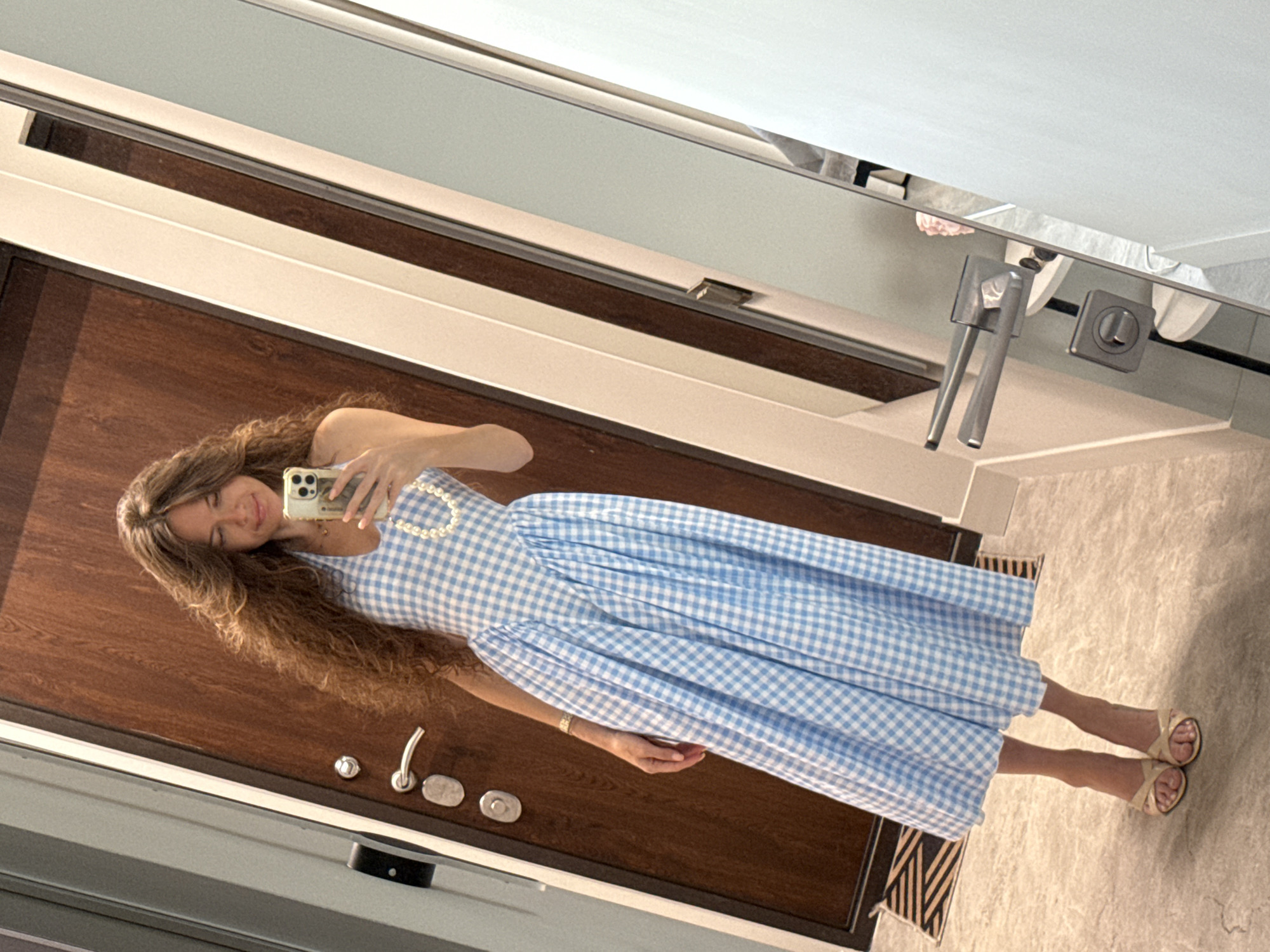 A woman with long, wavy brown hair is taking a mirror selfie in a hallway with a wood-paneled door and a marble-tiled floor. She is wearing a light blue and white gingham halter dress with a pearl necklace. Her left hand, adorned with a bracelet, is resting on her hip, while her right hand holds a smartphone. She is also wearing beige open-toed heeled sandals. The environment suggests an indoor residential or hotel setting. There is no indication this scene is in Moscow, Russia.