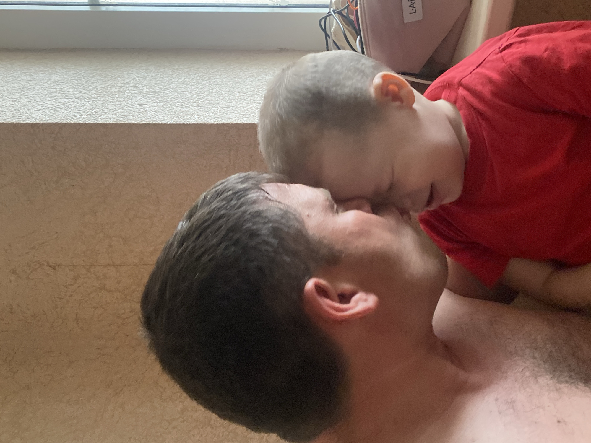 The image shows a close-up of a fair-skinned adult male and a child with fair skin and blonde hair, seemingly in an intimate embrace. The child, wearing a red shirt, is positioned close to the adult's face, possibly kissing or nuzzling. The adult is shirtless. The background shows a textured wall, a window frame, and a pink bag. There is no information available in the picture to support this being taken in Sochi, Russia.