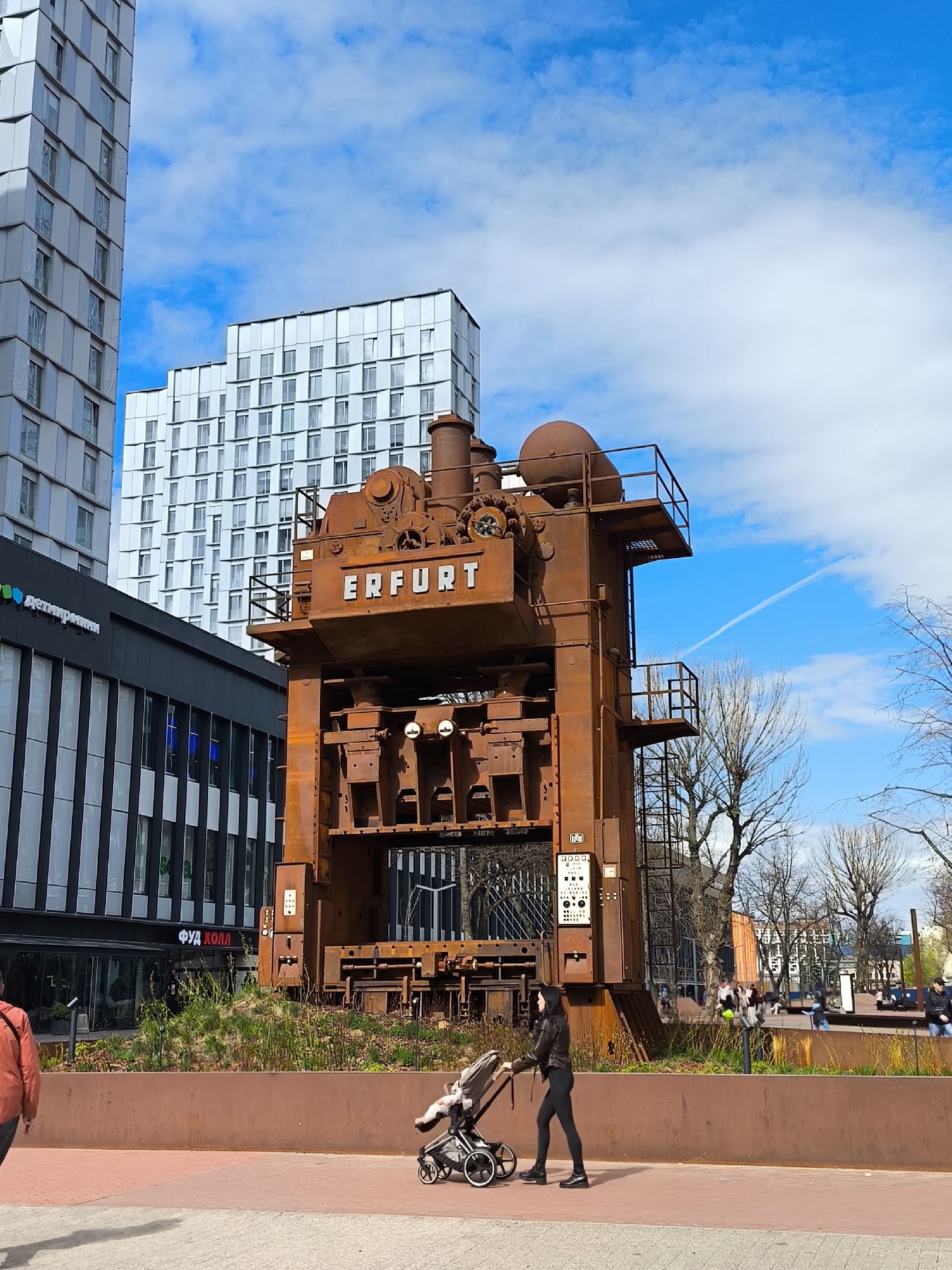A medium shot captures an open urban space in Moscow, Russia, featuring a massive, rusty industrial press prominently labeled "ERFURT" as its centerpiece. To the left, a modern building with alternating black and white vertical panels and a ground-level restaurant with signage in Cyrillic characters forms the backdrop. Behind the press, two taller, contemporary residential buildings rise, contrasting with the overcast sky partially obscured by scattered clouds and a contrail.
A woman in black activewear pushes a stroller on the paved walkway in the foreground. In the background, blurred figures of people strolling and relaxing on benches suggest a public gathering space. The press is situated within a raised planter bed filled with greenery, demarcating it from the pedestrian area. The overall lighting suggests daylight conditions.