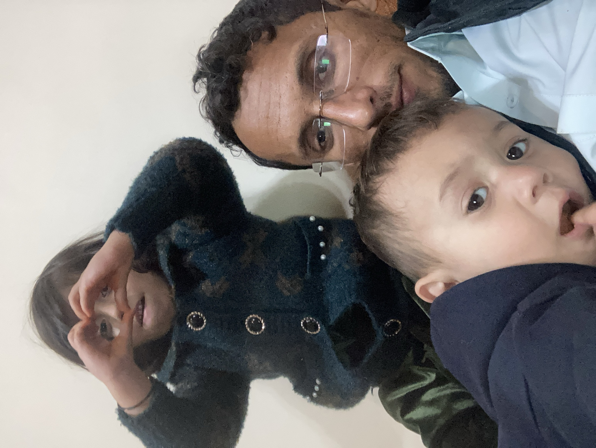 The image shows a selfie of a man with two children. The man is wearing glasses and a white shirt. One child is making a heart shape with their hands. The other child is looking directly at the camera. The location may be in Quetta City Tehsil, Pakistan.