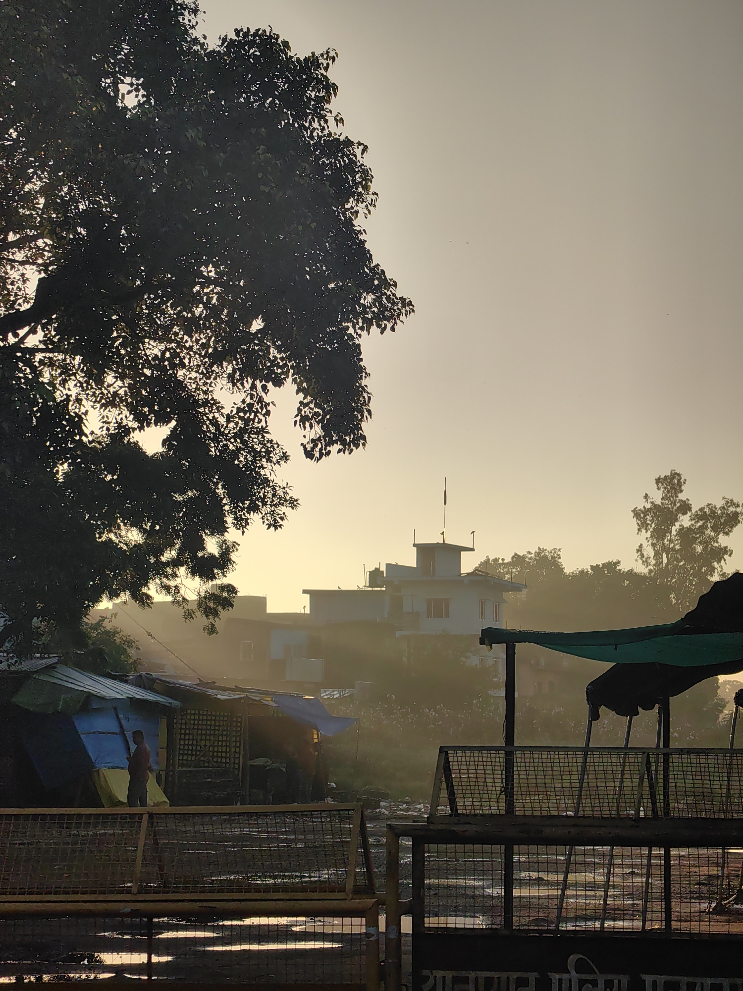 A low-angle, medium shot captures a hazy morning scene in Belahiya, Nepal. The foreground features a yellow metal barrier with horizontal bars and wire mesh, separating the viewer from a muddy, waterlogged area reflecting the soft morning light. Behind the barrier, several temporary structures with corrugated metal and tarpaulin roofs are visible, appearing to be makeshift shops or stalls. A single person, dressed in a red shirt and dark pants, stands within the frame of one of these structures, holding a long pole. Further back, a two-story building with a white facade and red-trimmed windows is partially obscured by mist, as are more distant trees. The sky is a pale, washed-out yellow, suggesting sunrise or sunset. The overall atmosphere is subdued and slightly damp, with a visible atmospheric haze that softens the details of the background.