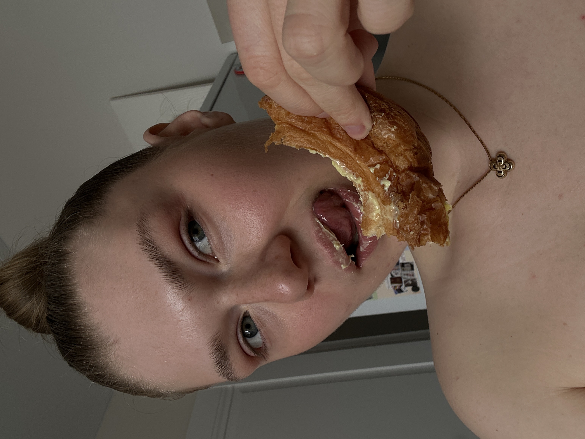 A person is eating a pastry, possibly a croissant, with a filling that appears to be cream or butter. The individual is fair-skinned with light hair pulled back into a bun. They are wearing a gold necklace with a clover-shaped pendant. The background shows a glimpse of a refrigerator. The image is shot at close range, likely by the person themselves. There is no indication this photo was taken in Kazan, Russia.