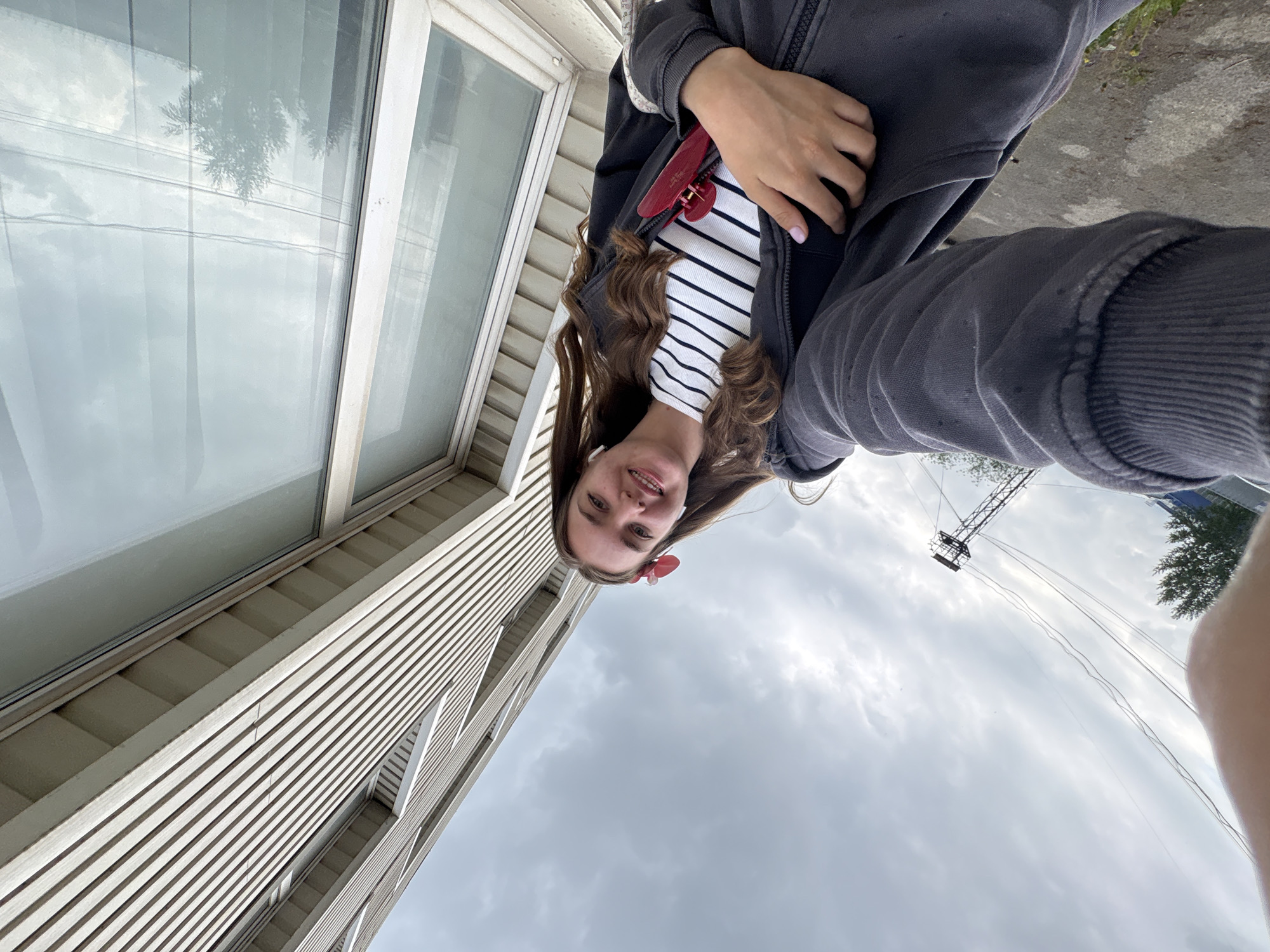A young woman is pictured in an upside-down selfie, likely taken in Perm, Russia, based on visual cues of architectural style and potential landmarks. She wears a striped shirt under a dark jacket and has a red hair accessory. She stands next to a building with a white facade and large windows. The sky is overcast. Overhead power lines are visible in the distance.