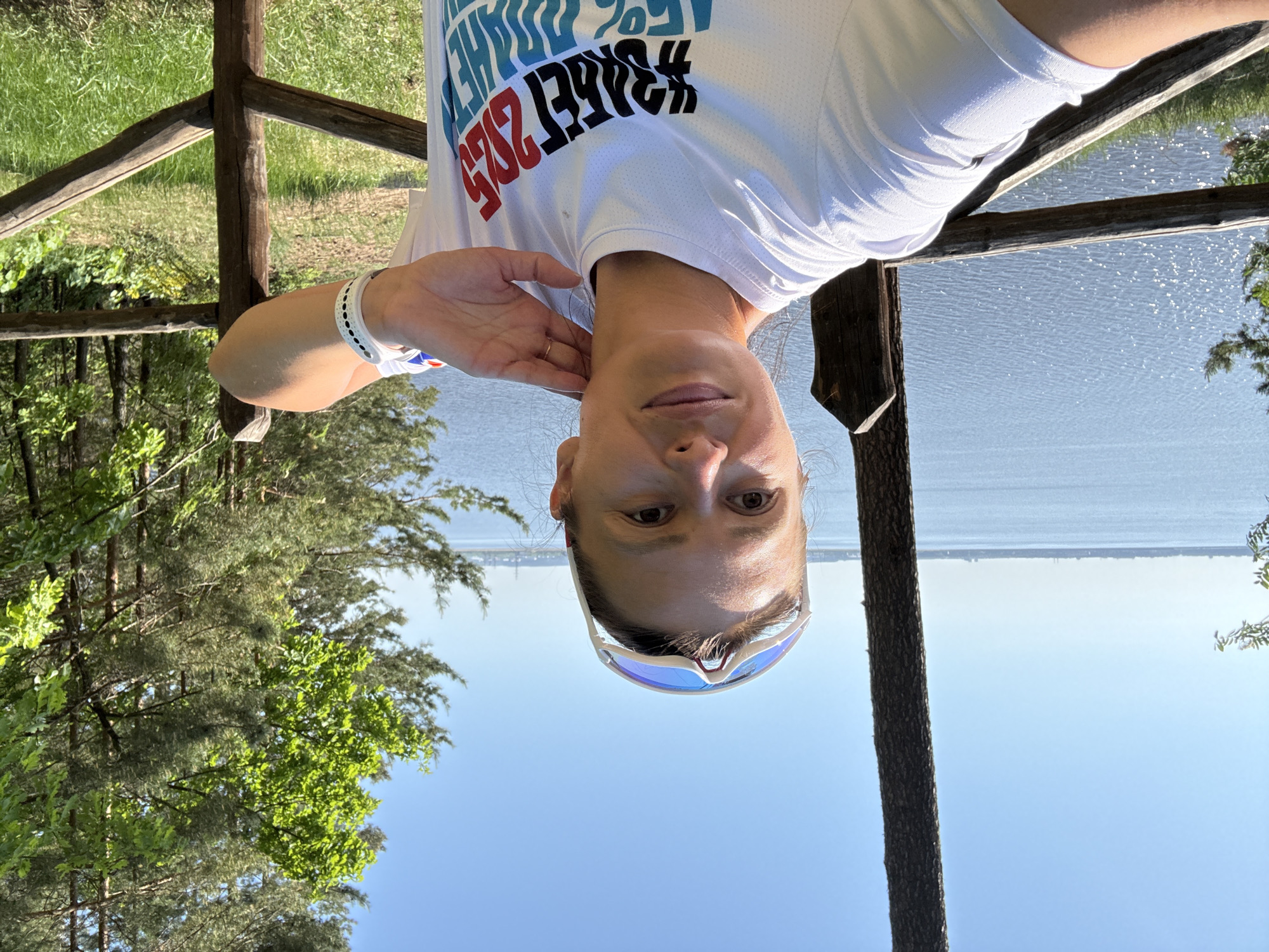 A woman is centered in the frame, wearing a white shirt with writing, with sunglasses on her head. She has fair skin and her hand is near her ear. She stands near a wooden structure. Behind her is a view of a body of water and trees. The image is taken outdoors in Noviy, Russia, during the day.