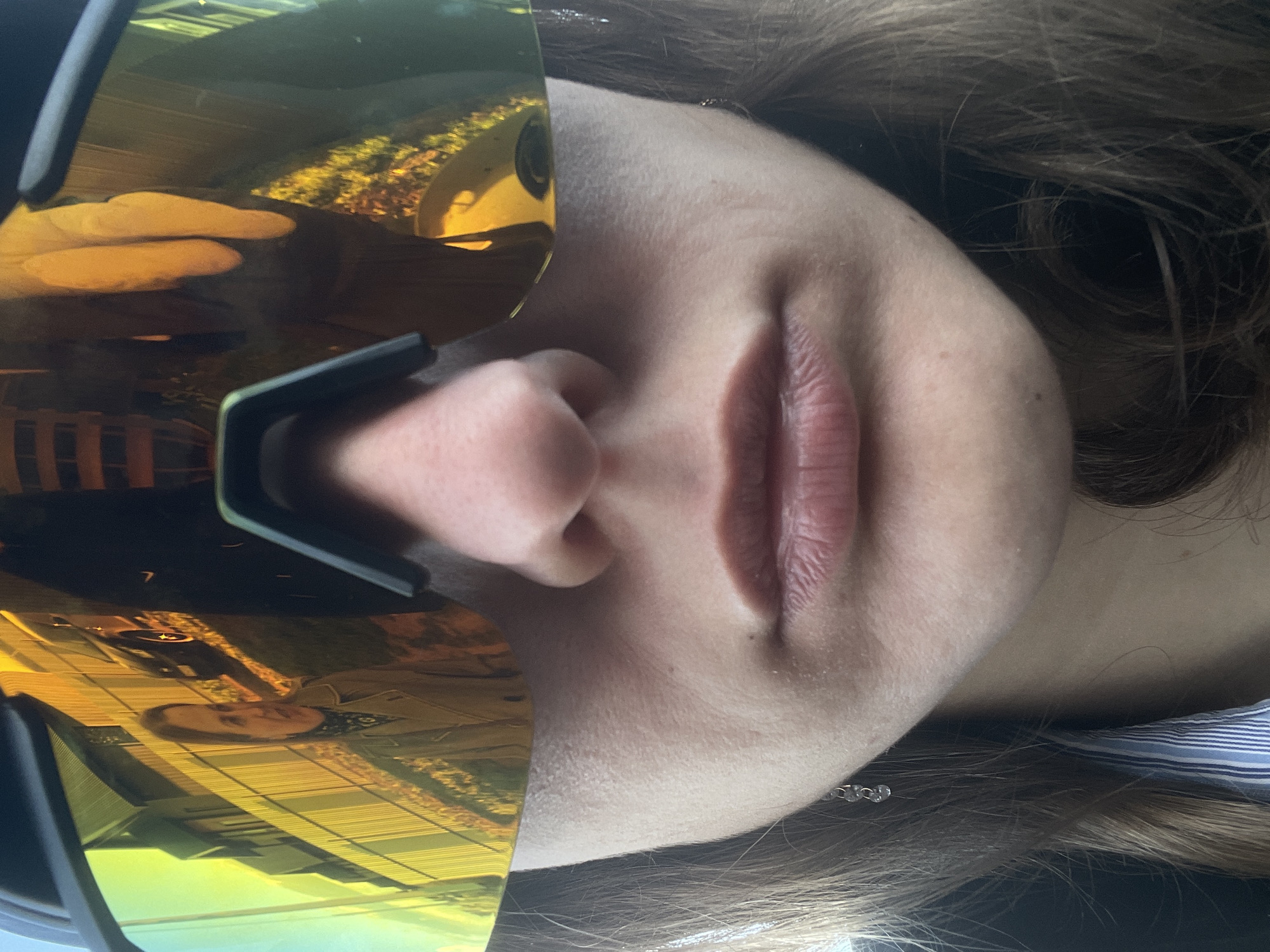 The image depicts a close-up view of a person's face, partially obscured by reflective eyewear. The skin is fair, with visible lips and a nose. The eyewear reflects an external scene, including structures with horizontal lines and what appears to be foliage. The person has light brown hair and is wearing a garment with blue and white stripes. The image does not provide enough information to determine the location within Подшивалово, Russia, or any specific actions or interactions.