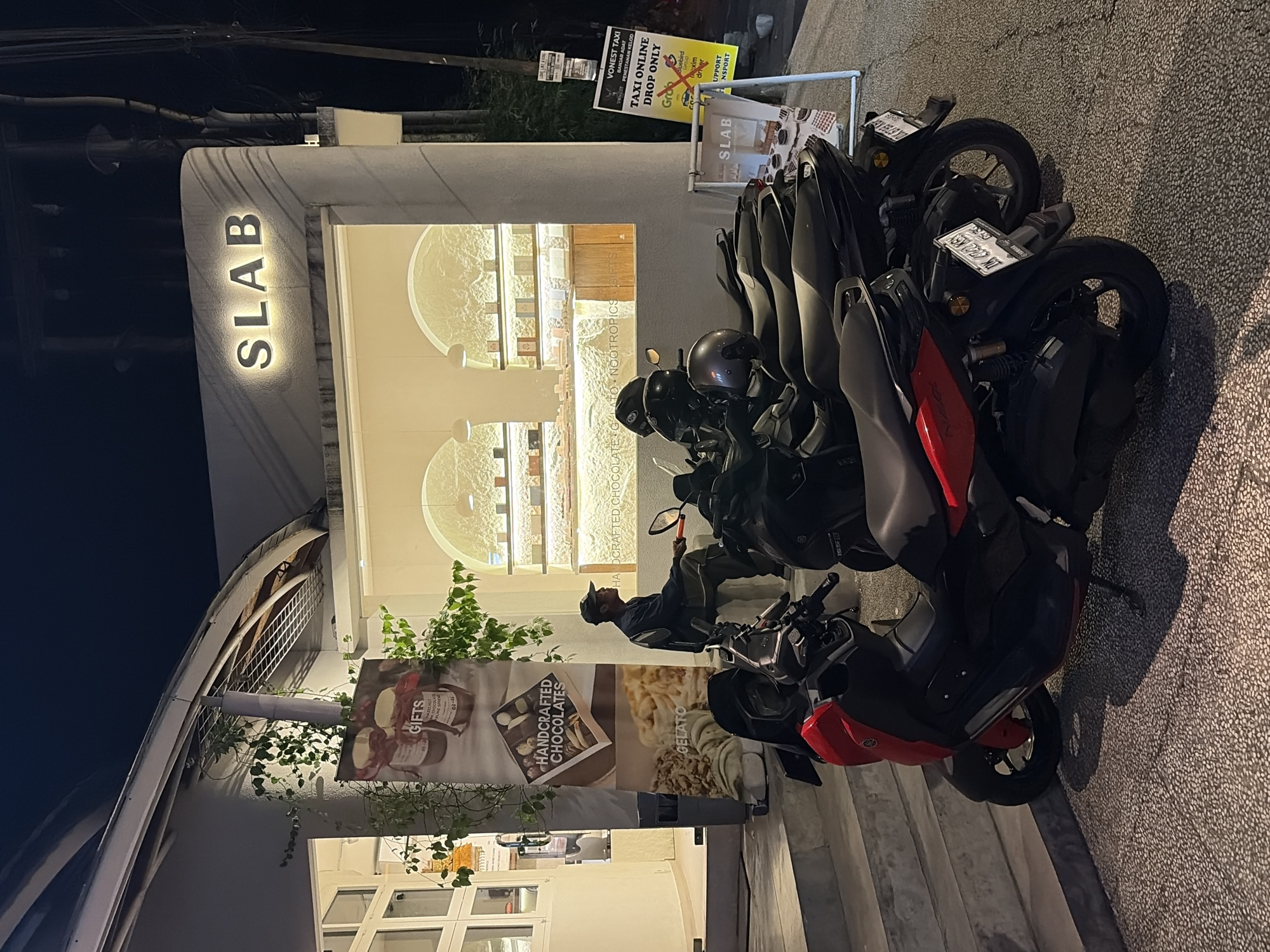 A man wearing a dark cap and a dark jacket sits on a low stool outside a shop named "SLAB" in Ubud District, Indonesia. The shop front is illuminated, displaying shelves filled with goods. Several motorcycles are parked on the pavement in front of the shop, with a red and black scooter in the foreground. A banner displaying "GIFTS" and "HANDCRAFTED CHOCOLATES" is visible on the left side of the entrance. A yellow sign for "VONEST TAXI" is placed near the motorcycles. The scene is lit by artificial lights and the darkness of night.