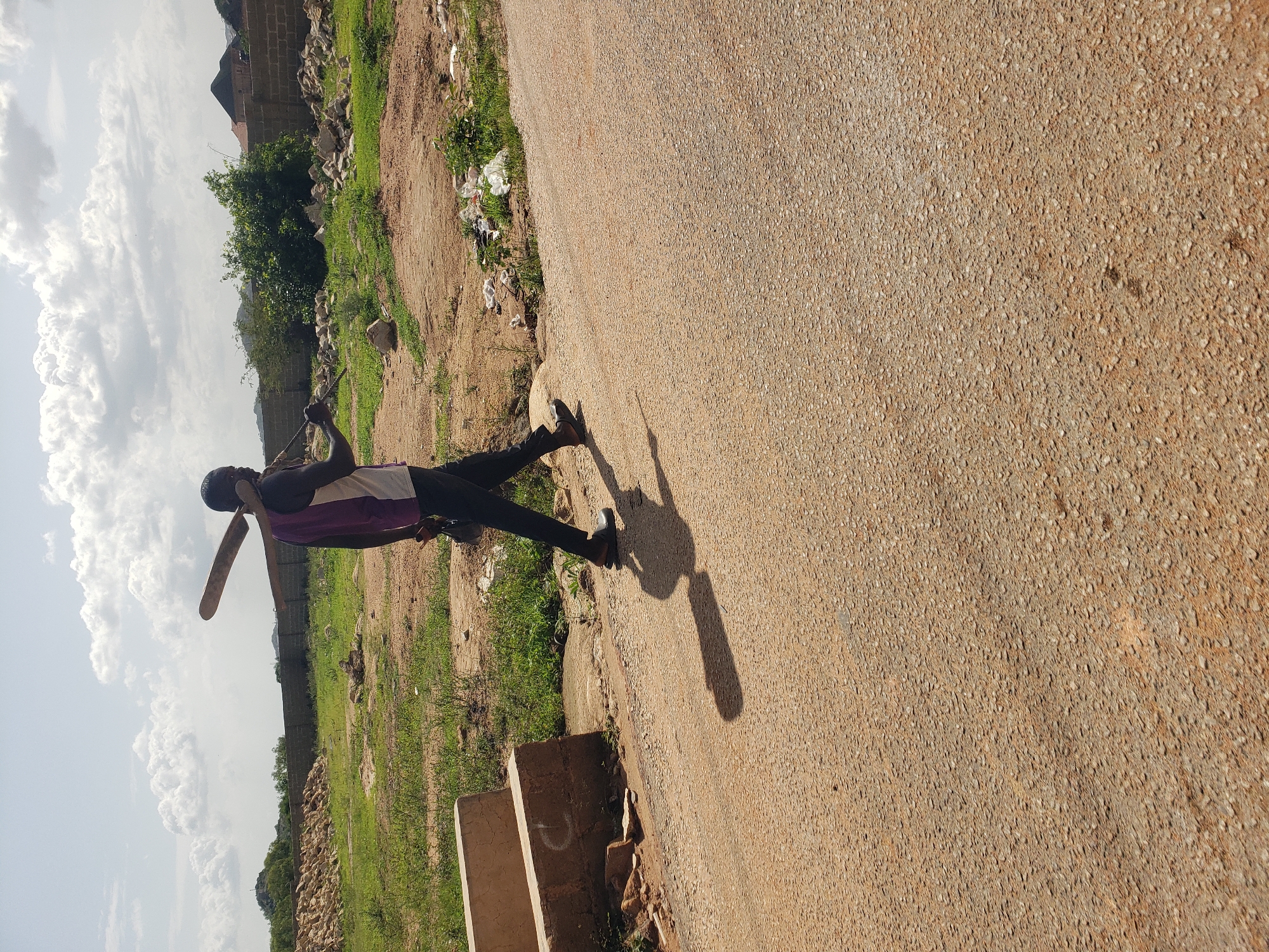 A full-shot, eye-level, outdoor, daylight, candid photograph captures a Black man walking on an asphalt road in Gura Top, Nigeria. The man is walking from left to right, facing away from the viewer, and is carrying a wooden object, possibly a paddle, over his right shoulder. He is wearing a purple and white tank top, dark pants, and dark footwear. His shadow is cast on the road. The background features a grassy field with scattered rocks, a brick wall, trees, and buildings. The sky is cloudy with some sunlight breaking through. The scene appears to be an undeveloped area with construction materials and debris visible.