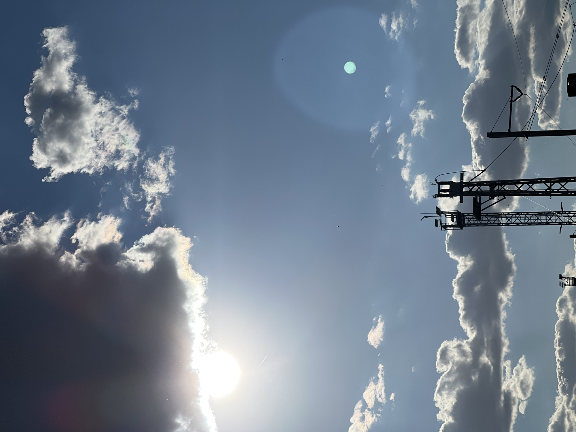 The image captures a sky scene in Újezd u Brna, Czechia. Silhouetted against a bright sky filled with scattered clouds, several structures are visible. In the foreground, two tall, dark, lattice-like towers stand, one on the left and another partially visible on the right. Power lines crisscross the frame between these structures. The sky is a mix of bright blue and areas obscured by clouds. A prominent, bright sun is visible in the upper left quadrant, with a lens flare creating a luminous effect and a faint contrail from an aircraft. In the upper right quadrant, a distinct, teal-colored circular artifact is present, possibly a reflection or lens anomaly. The clouds are varied in density, with some appearing dark and heavy, while others are lighter and wispy, catching the sunlight. The overall impression is of an outdoor scene, possibly taken from an elevated position, with a focus on the sky and man-made infrastructure.