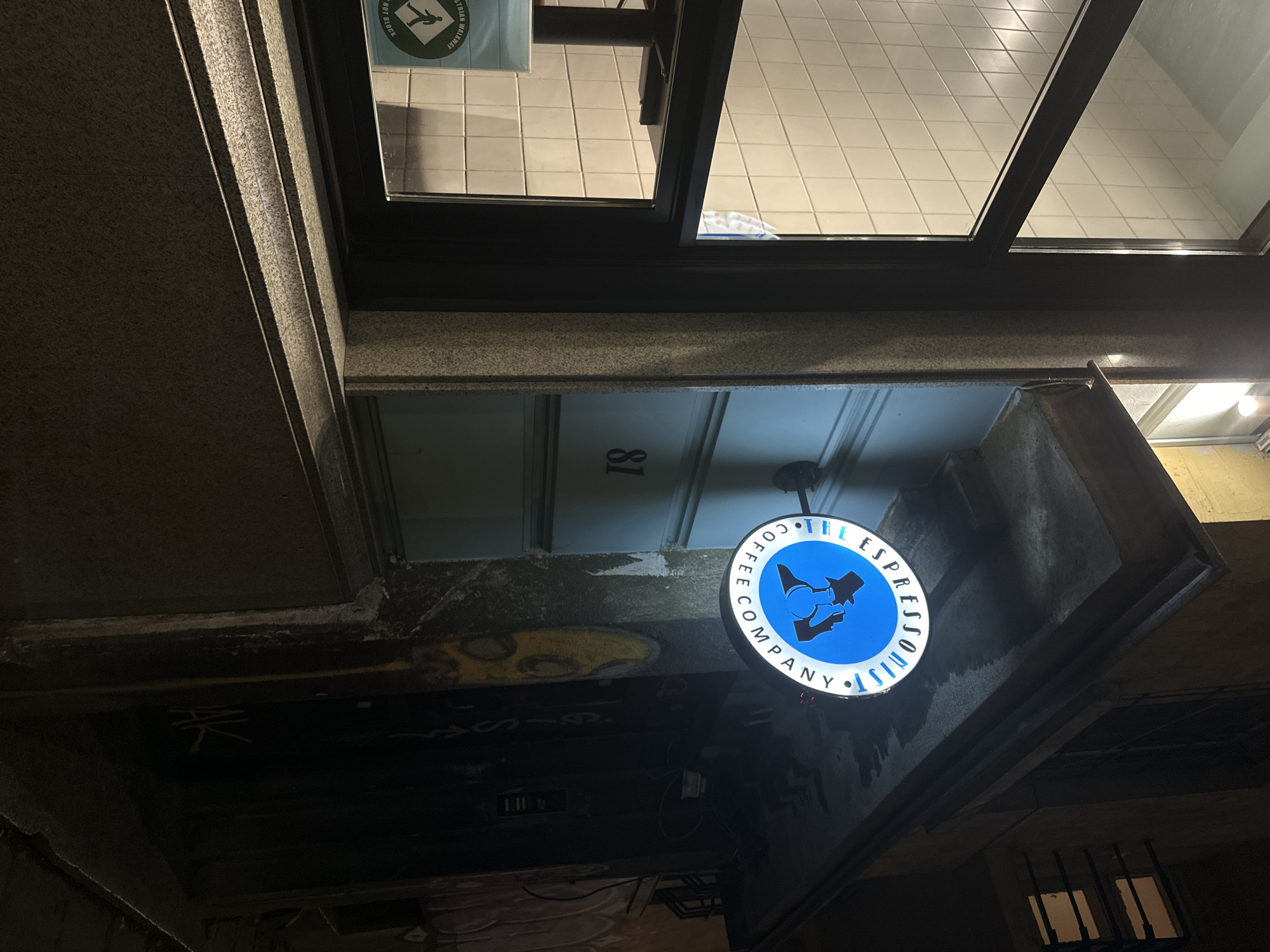 The image shows the exterior of "The Espressoist Coffee Company" located at 81 in Sofia, Bulgaria. The sign is circular, blue with white lettering and depicts a stylized figure holding an espresso pot. The sign is mounted under a concrete overhang, above an area with graffiti. Above the overhang is a window with multiple square panes. The surrounding architecture is grey and appears to be stone or concrete. The scene is dimly lit.