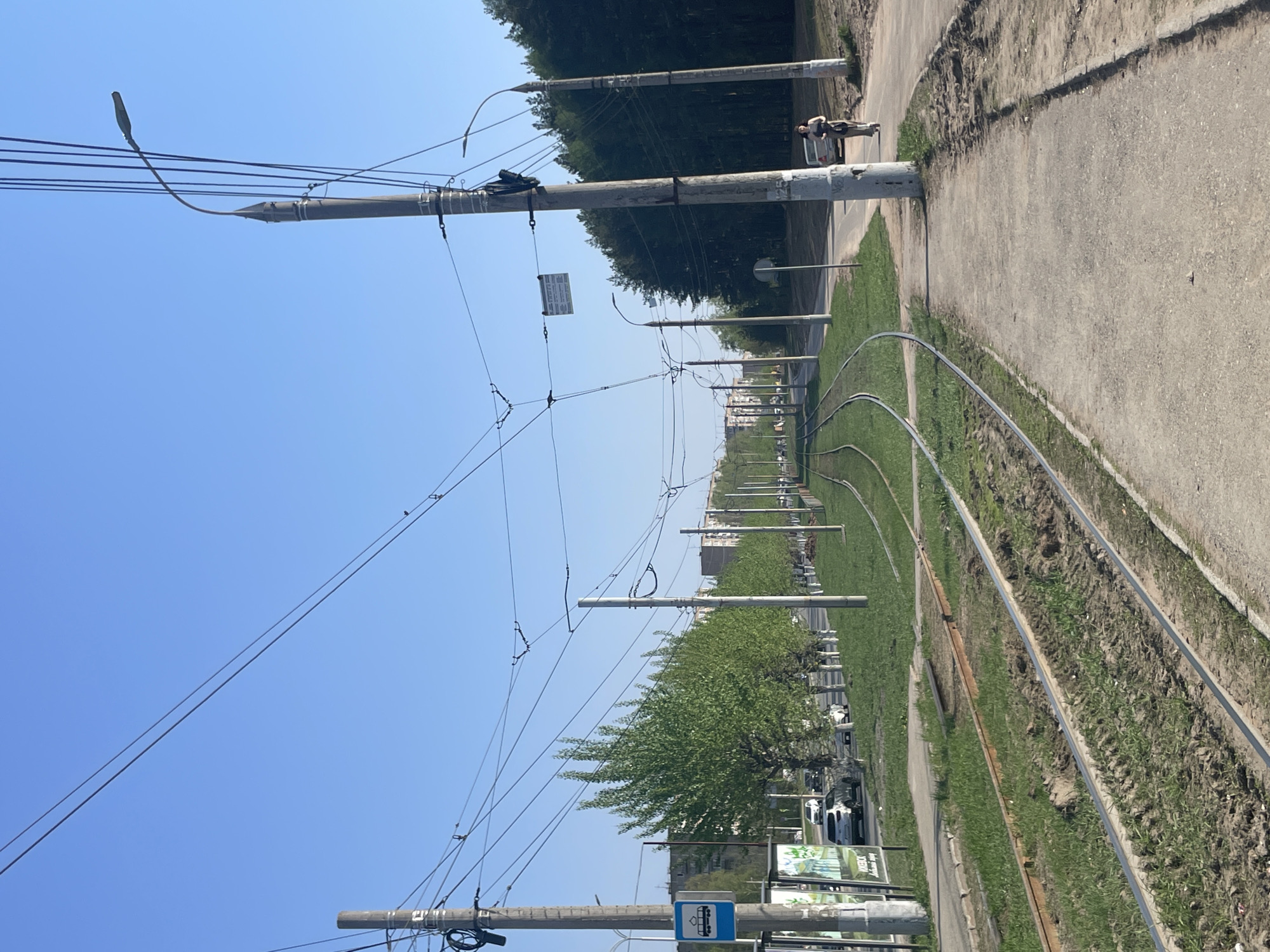 The image depicts a street scene in Izhevsk, Russia. A light-blue sky is visible above a row of utility poles with numerous overhead wires. The street features tram tracks running along a grassy median. A sidewalk runs alongside the median. A cyclist and a pedestrian are visible on the sidewalk. Cars are parked along the side of the street behind the median. A bus stop sign is visible in the foreground. Billboards also stand along the road. The background shows buildings and trees lining the horizon.