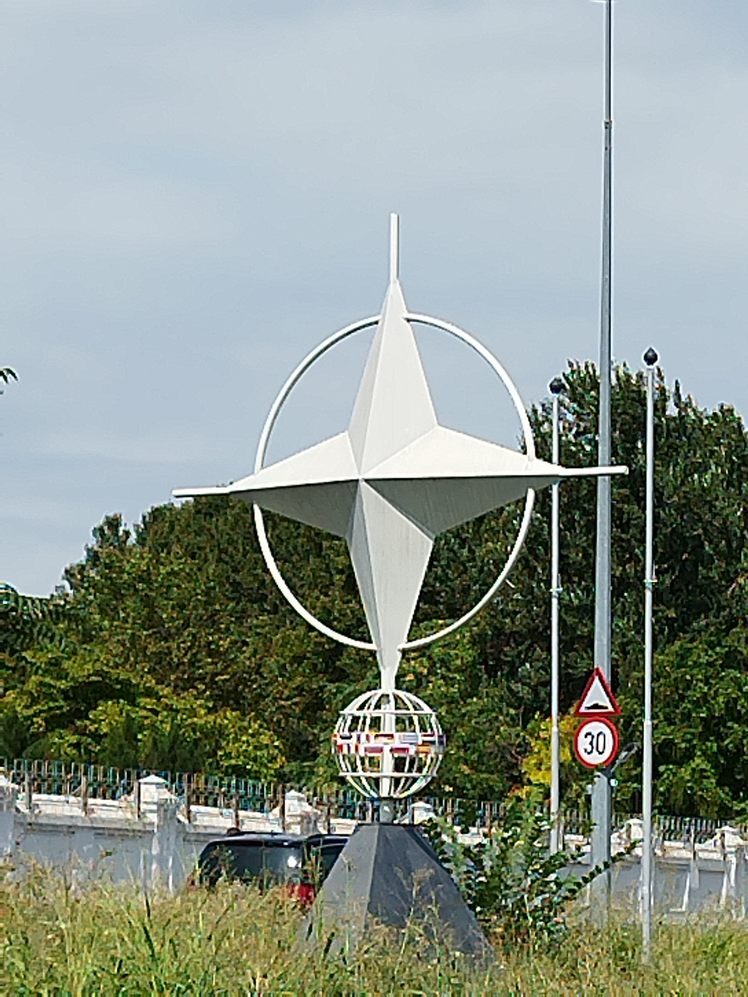 The image shows a monument that appears to be the NATO star, a prominent symbol of the North Atlantic Treaty Organization. The monument is located outdoors, in what seems to be a public space with trees and foliage in the background. A speed limit sign indicating 30 is visible on a pole to the right of the monument. A black car is partially visible behind a white fence. The monument itself consists of a large, four-pointed star inside a circle, mounted on a pedestal that includes a spherical structure. The overall scene suggests an urban or suburban setting.