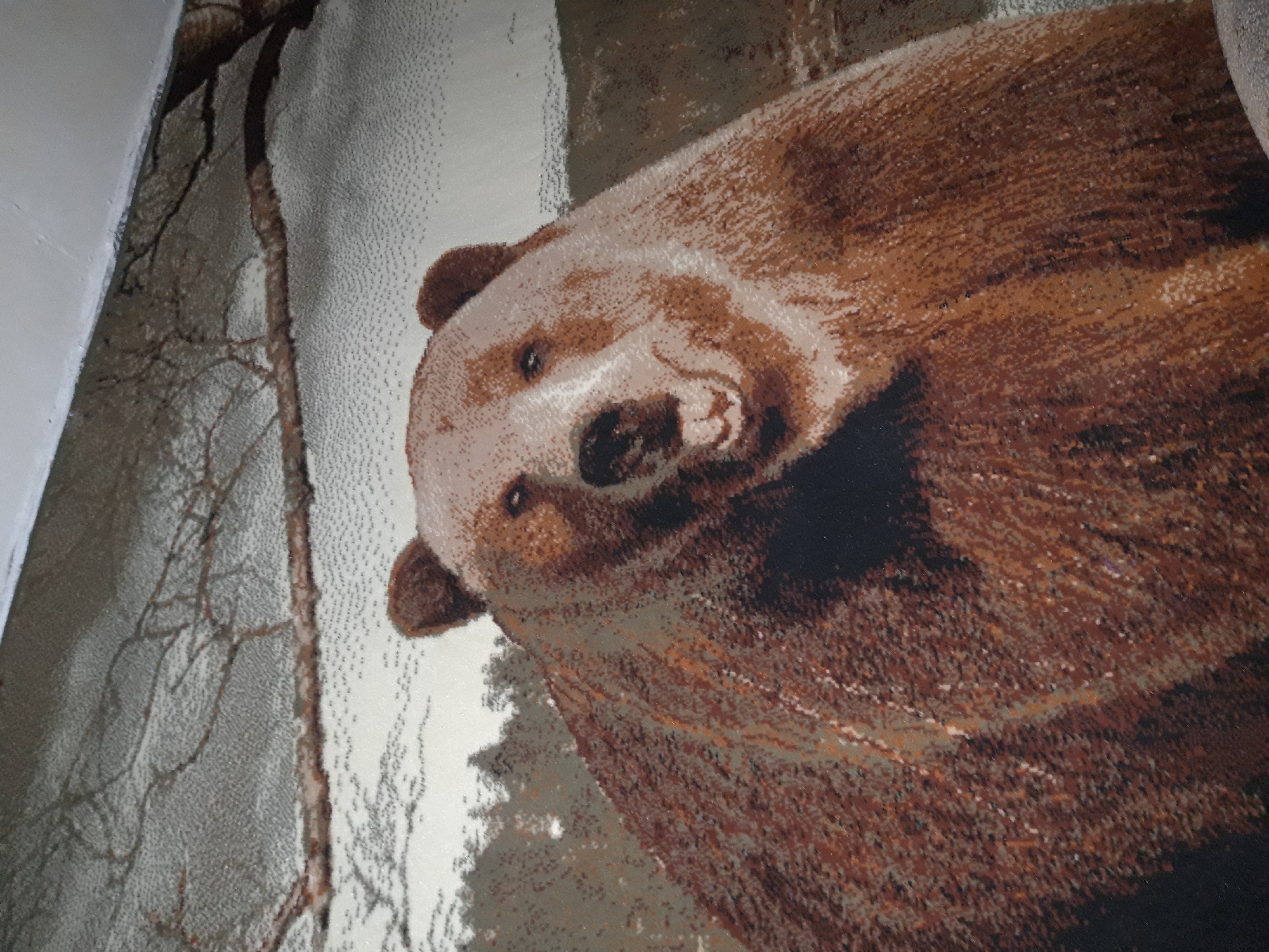 The image displays a close-up of a tapestry or rug featuring a brown bear in a natural setting. The bear's head and upper body dominate the frame, with its fur rendered in varying shades of brown and white. The background depicts a forest or wooded area with bare branches visible in the upper portion of the image. The tapestry appears to be hung on a wall, indicated by a white ceiling line at the top. The overall impression is of a detailed and realistic depiction of a bear. There are no people or specific locations within the city of Макаров ауылдық округі, Kazakhstan, visible in this image.