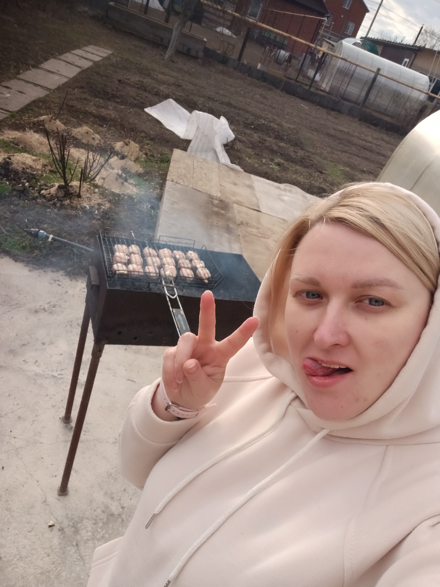 A blonde-haired woman, wearing a light-colored hooded sweatshirt, is depicted in a selfie outdoors. She faces the camera, making a peace sign with her left hand and playfully sticking her tongue out. On her left wrist, a pink wristband or watch is visible.
Behind her, sausages are actively grilling on a dark metal mangal (barbecue grill), emitting smoke. The setting appears to be a backyard or garden, likely in Chaikovsky, Russia, as suggested by the context. The ground consists of bare earth patches and concrete or paved sections. Several small bushes or plants are visible, mulched with straw or wood shavings.
In the mid-ground, there's a rustic wooden structure and white plastic sheeting. Further back, residential houses, fences, and multiple greenhouses or hothouses can be observed. The sky is overcast, providing soft, diffused lighting, indicative of daytime. The bare ground and hooded clothing suggest either early spring or late autumn. The woman's expression and gesture convey a casual and light-hearted mood. Faint, largely illegible text, possibly "INOX," is visible on the metal handle of the grill rack.