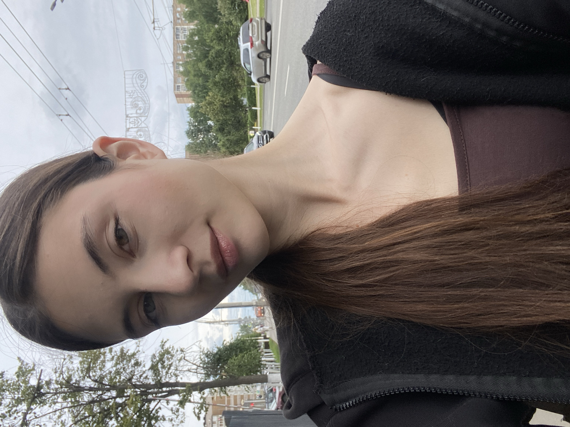 The image is a portrait of a young woman taken outdoors in Izhevsk, Russia. She is turned slightly towards the camera, with her head tilted downwards. Her long, dark brown hair cascades down her right shoulder. She is wearing a dark brown top and a black zip-up jacket. Her expression is neutral, with a slight hint of a smile.
The background shows a street scene with cars and trees. The sky is overcast, suggesting it is daytime with cloudy weather. There are power lines visible in the upper left portion of the image, and a building can be seen in the distance. The overall setting appears to be an urban environment. There is no visible text in the image. The camera angle is from a low perspective, looking up at the woman.