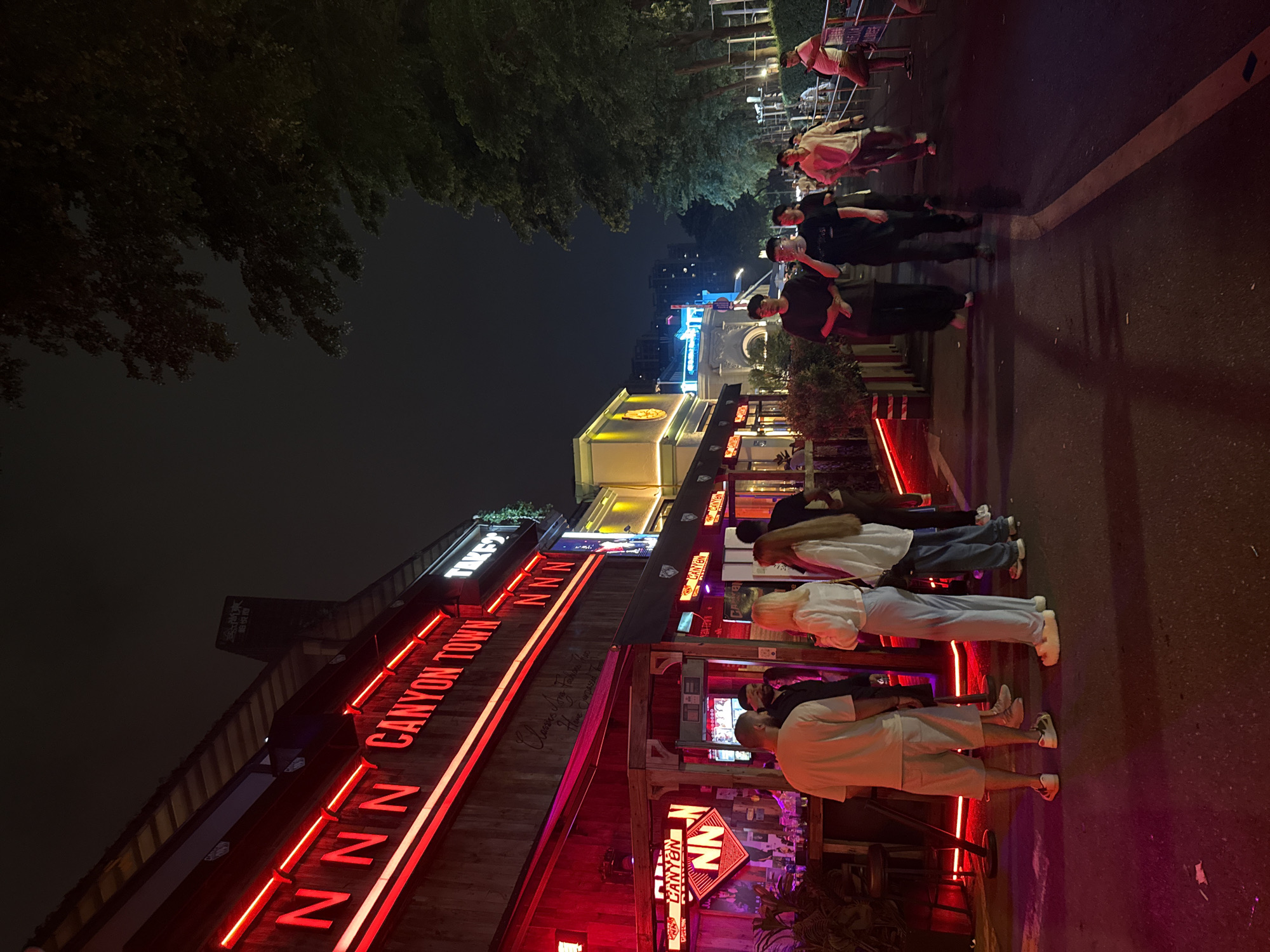 The image depicts a night scene in Tianhe District, China. Several pedestrians walk along a brightly lit street near a commercial building. The building is adorned with neon signs, including "CANYON TOWN" in prominent red lettering. Additional lighting casts a red hue on the street and surrounding area. The image includes partial views of trees and other buildings in the background.