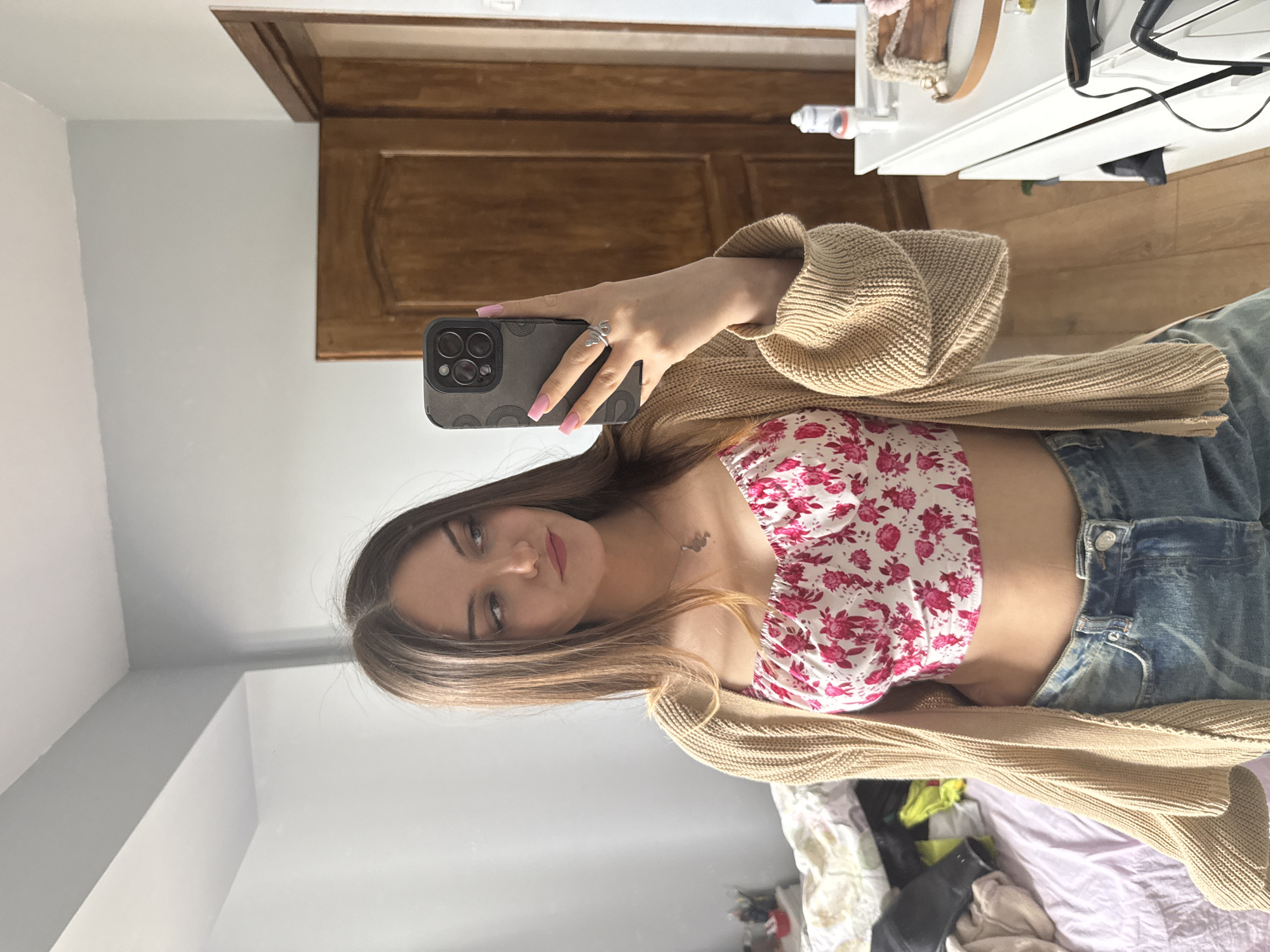 A young woman is taking a selfie in a room. She is holding a phone with a gray case and pink nail polish is visible on her fingers. The woman is wearing a floral crop top and a beige cardigan, paired with denim jeans. A silver butterfly ring is on her finger. The background includes a wooden door, a dresser with various objects on top, and a glimpse of a bed with clothing and other items scattered on it. The room is located in a city, however the exact location in the city of Sofia, Bulgaria cannot be deduced.