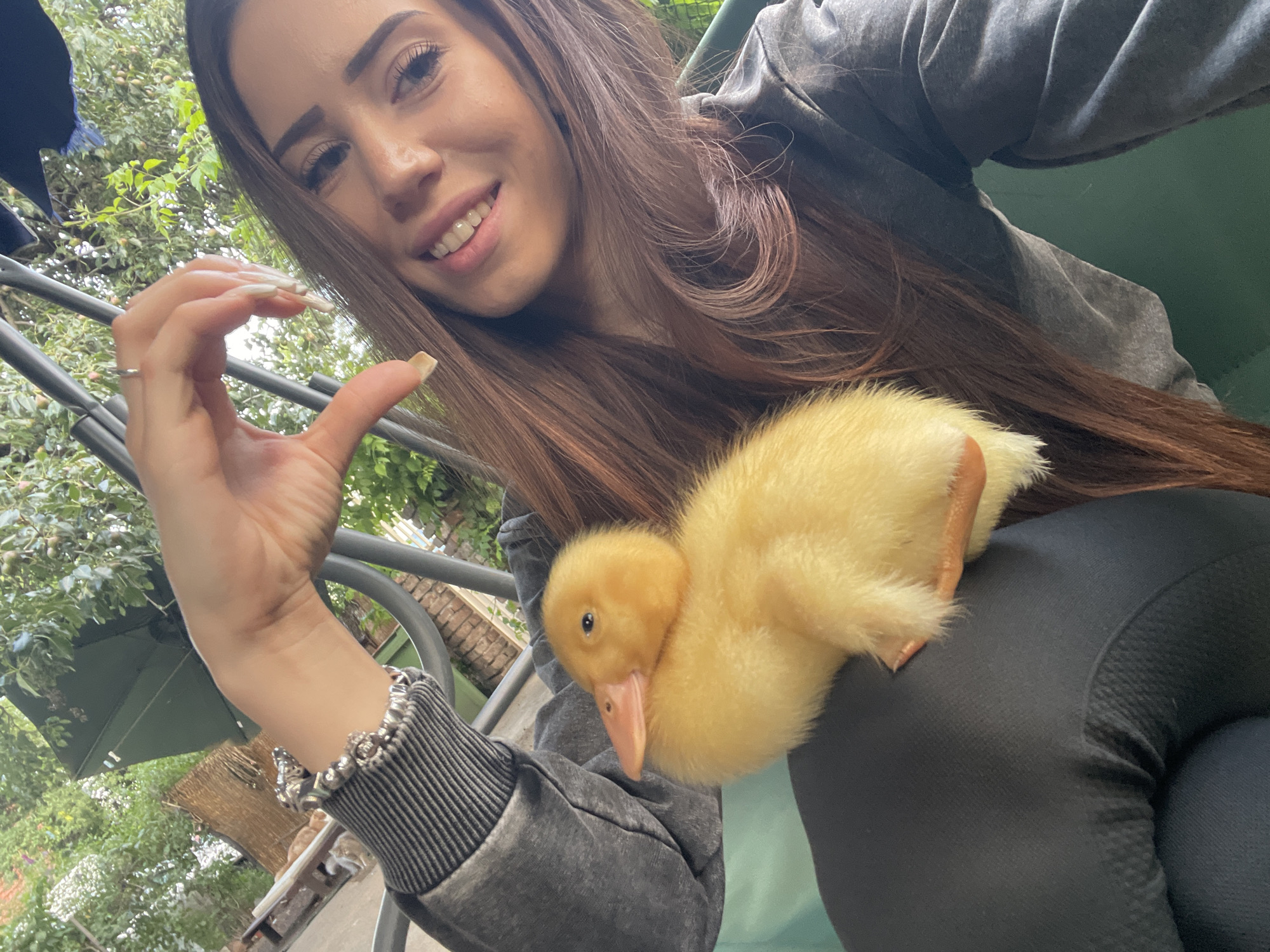In an outdoor setting, a young woman with long, brown hair makes a heart shape with her hand near her face, smiling. She is holding a fluffy yellow duckling on her lap. The woman is wearing a grey sweatshirt and a bracelet on her left wrist. The duckling is positioned on the left side of the frame, facing left. The background consists of lush green foliage and some wooden structures, suggesting a garden or backyard environment. The lighting indicates it is daytime.